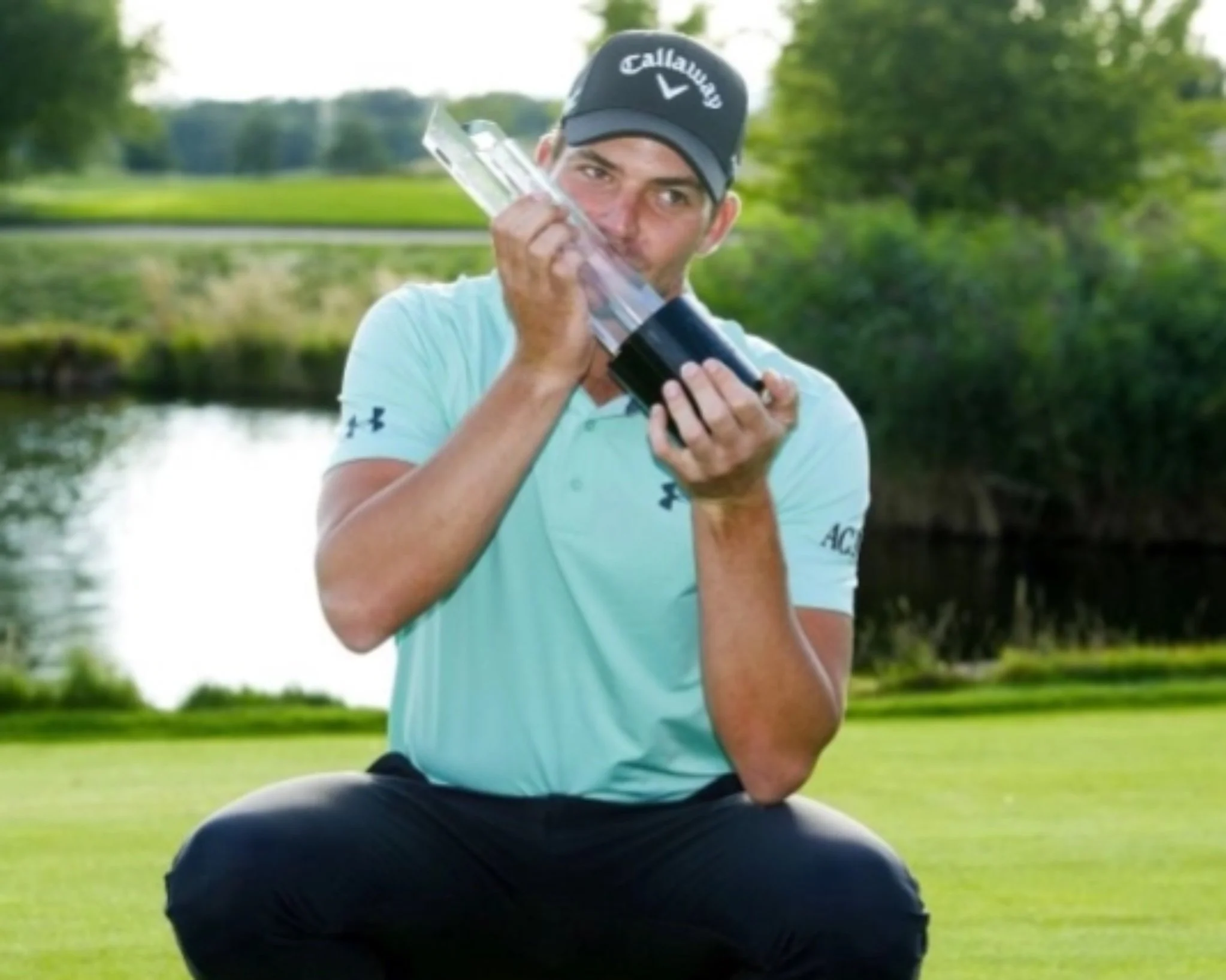 A man wearing a Callaway cap and light blue golf shirt is sitting outdoors on grass, holding and kissing a large golf trophy with a black base.