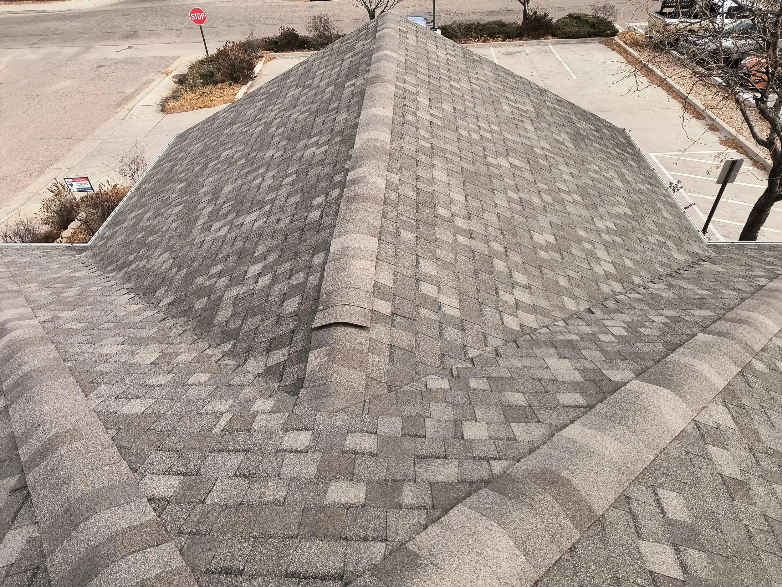 Aerial view of a gray shingled roof with a cross gable design, surrounded by a parking lot, sidewalk, and a stop sign.