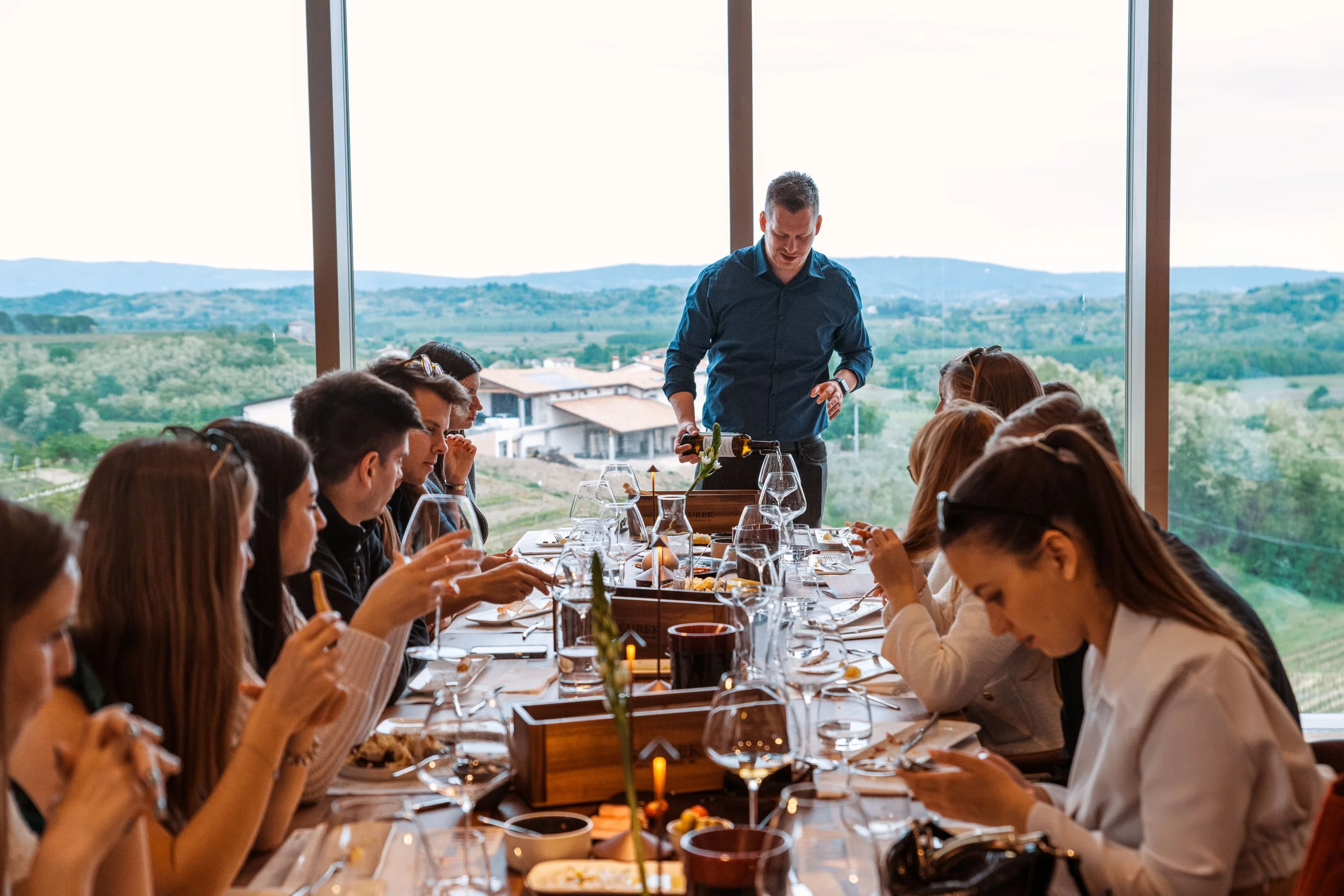 A group of people dining at a long table in a restaurant with large windows overlooking a scenic countryside.