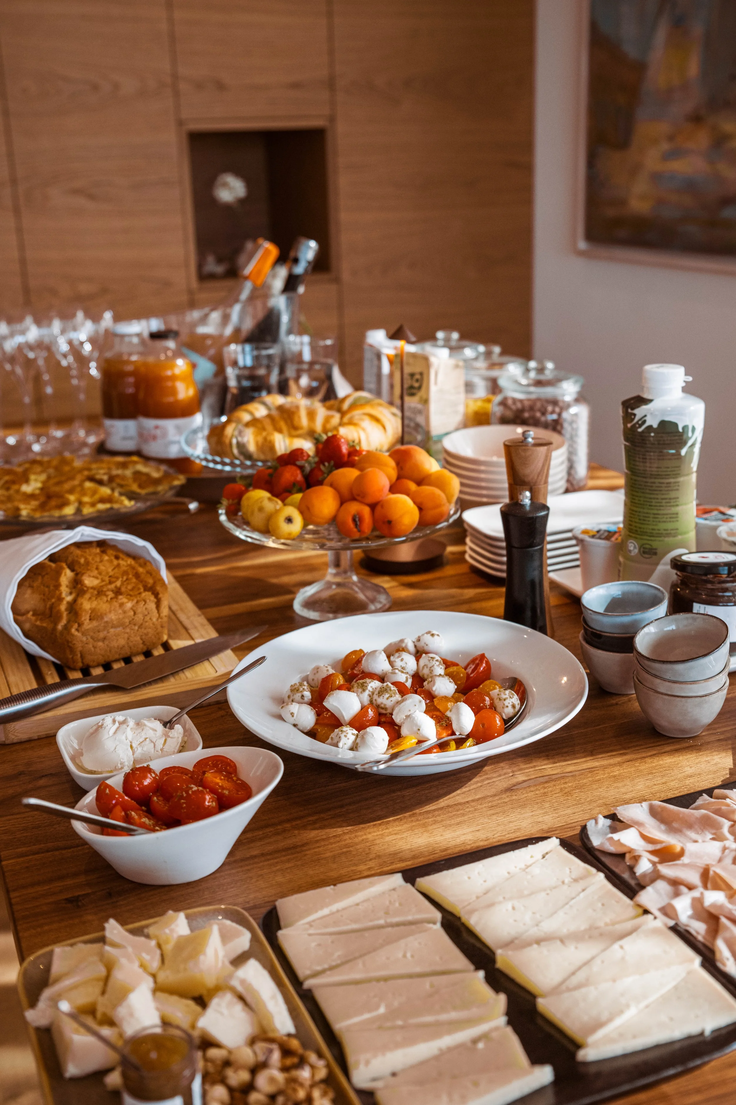 A breakfast spread on a wooden table with various foods including sliced cheese, cherry tomatoes, mozzarella salad, bread, pastries, small bowls of jam or honey, and jars of spreads or preserves.