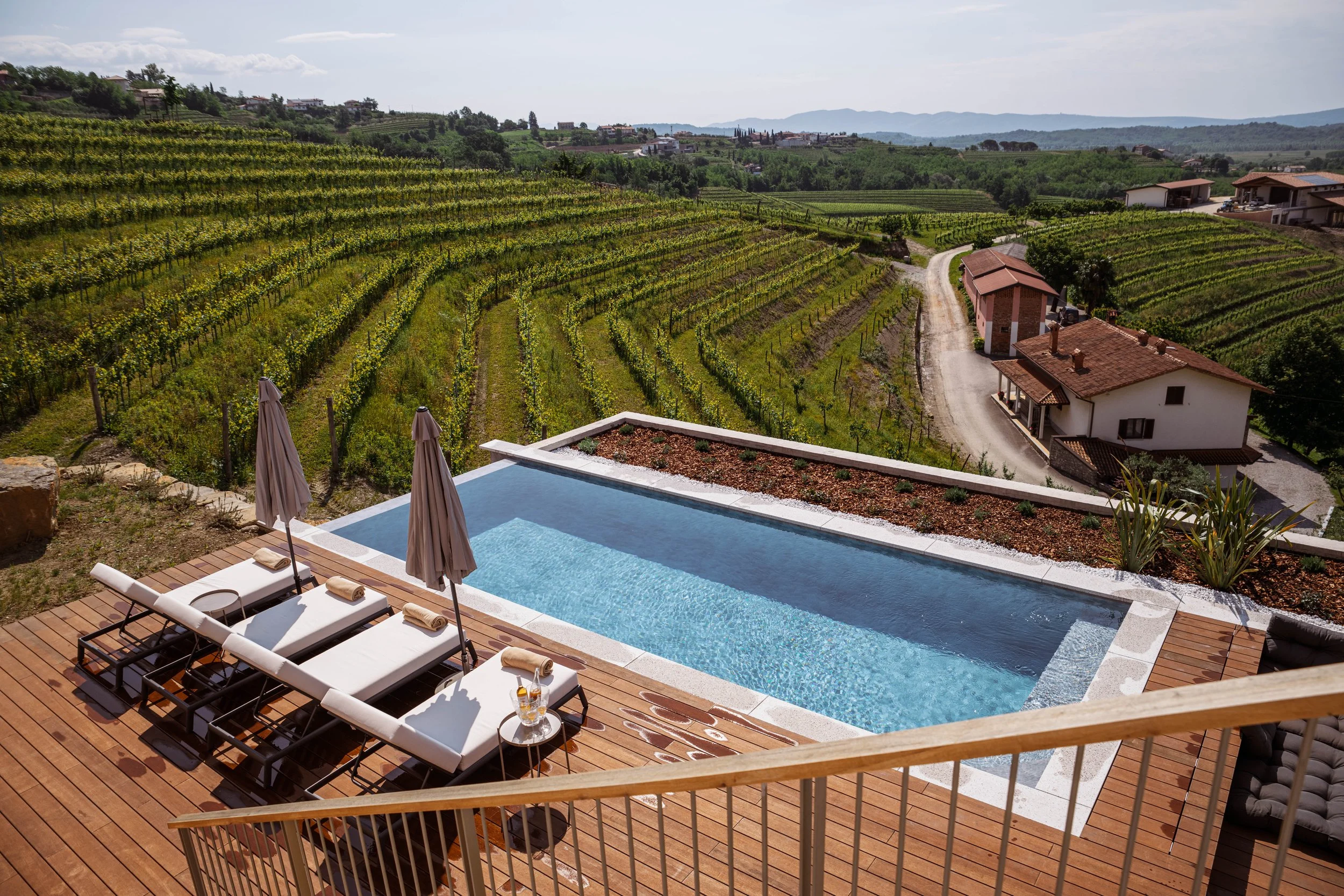 A swimming pool with lounge chairs on a wooden deck overlooking rolling hills with vineyards and houses.