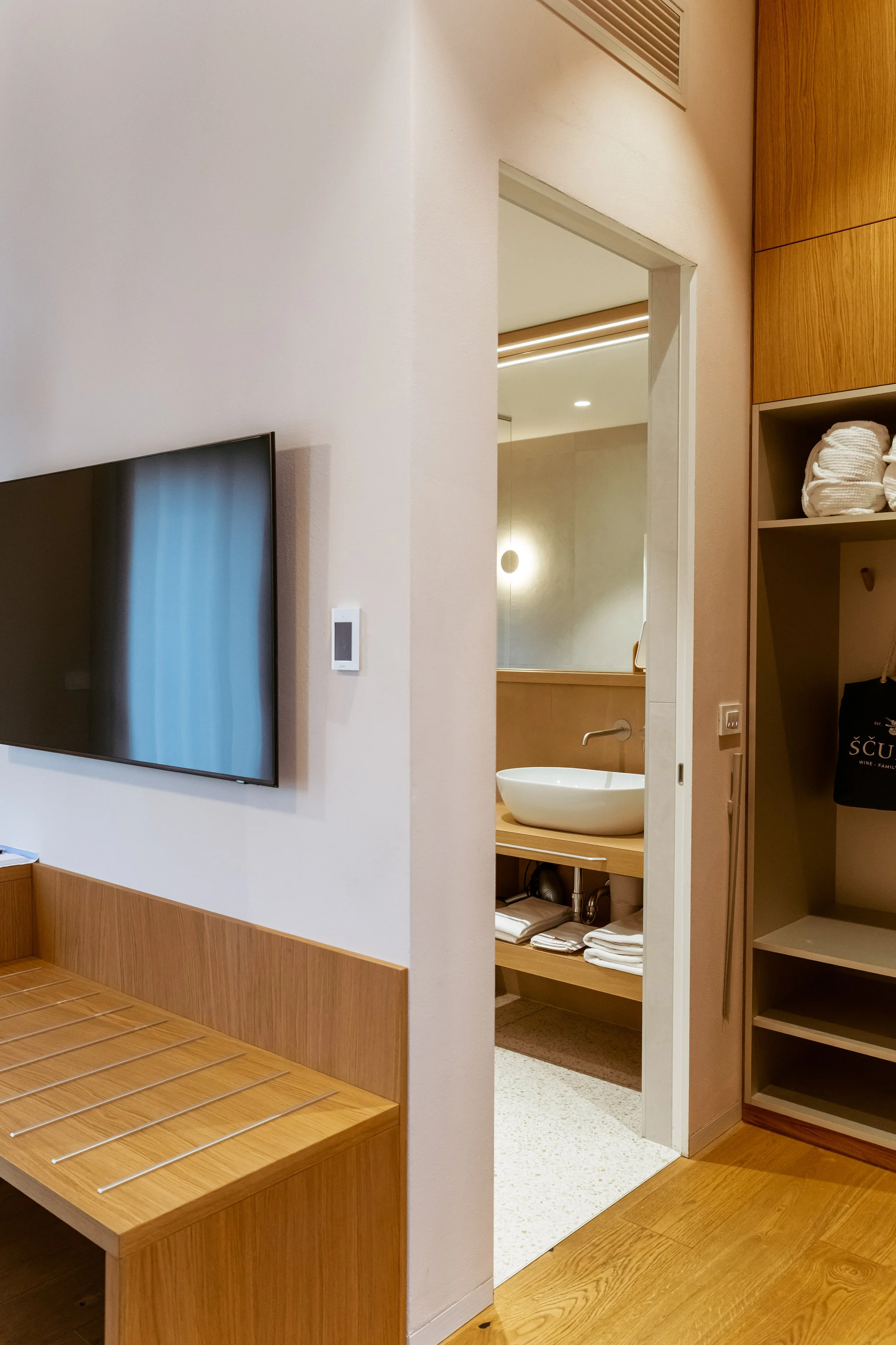Interior view of a hotel room with a flat screen TV on a white wall, a small bathroom with a vessel sink visible through an open door, and wooden furniture including shelves, a bench with metal rods, and a countertop with towels.