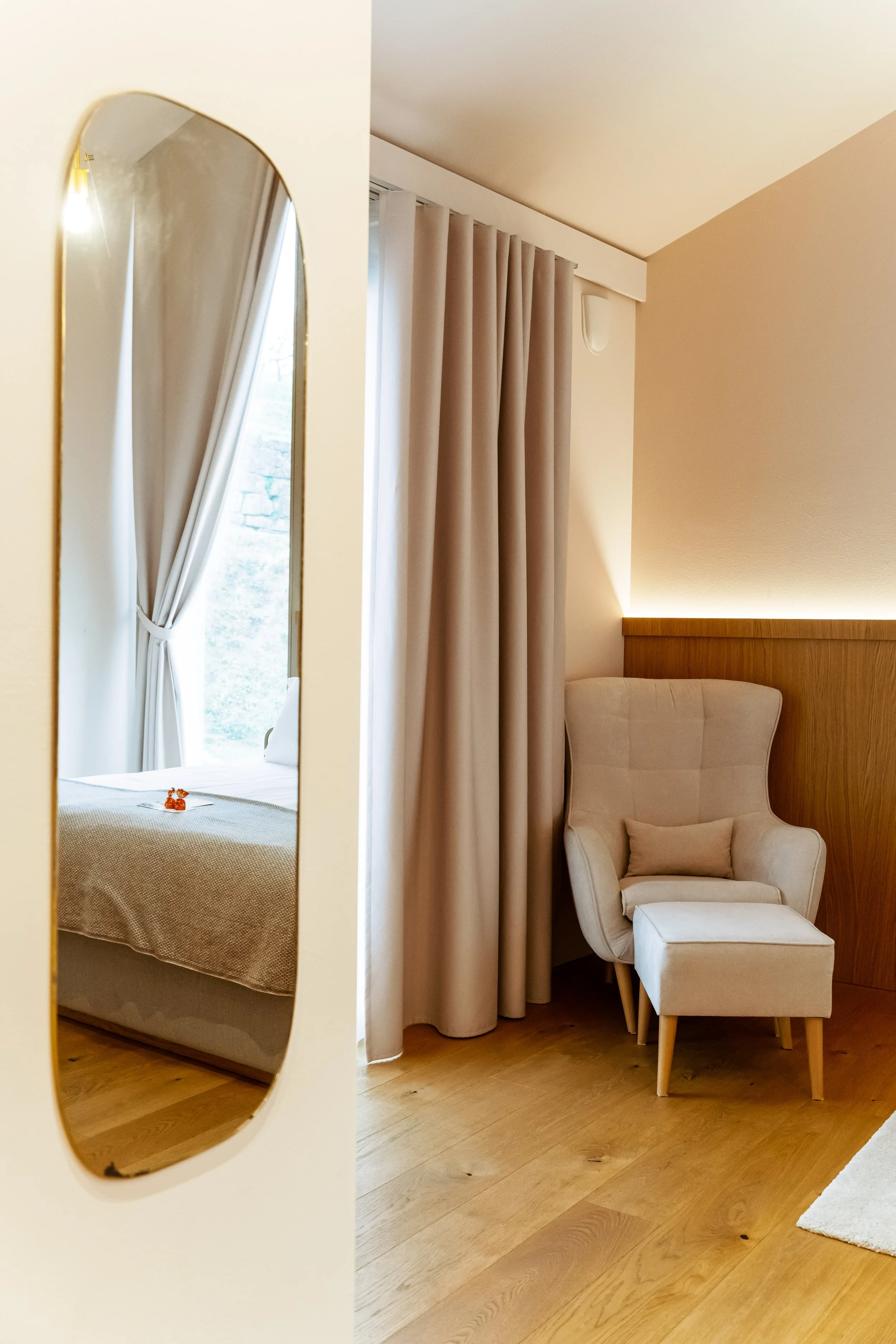 A cozy bedroom with beige curtains, a wooden headboard with LED lighting, and a white armchair with a matching cushion and ottoman, reflected in a full-length mirror.