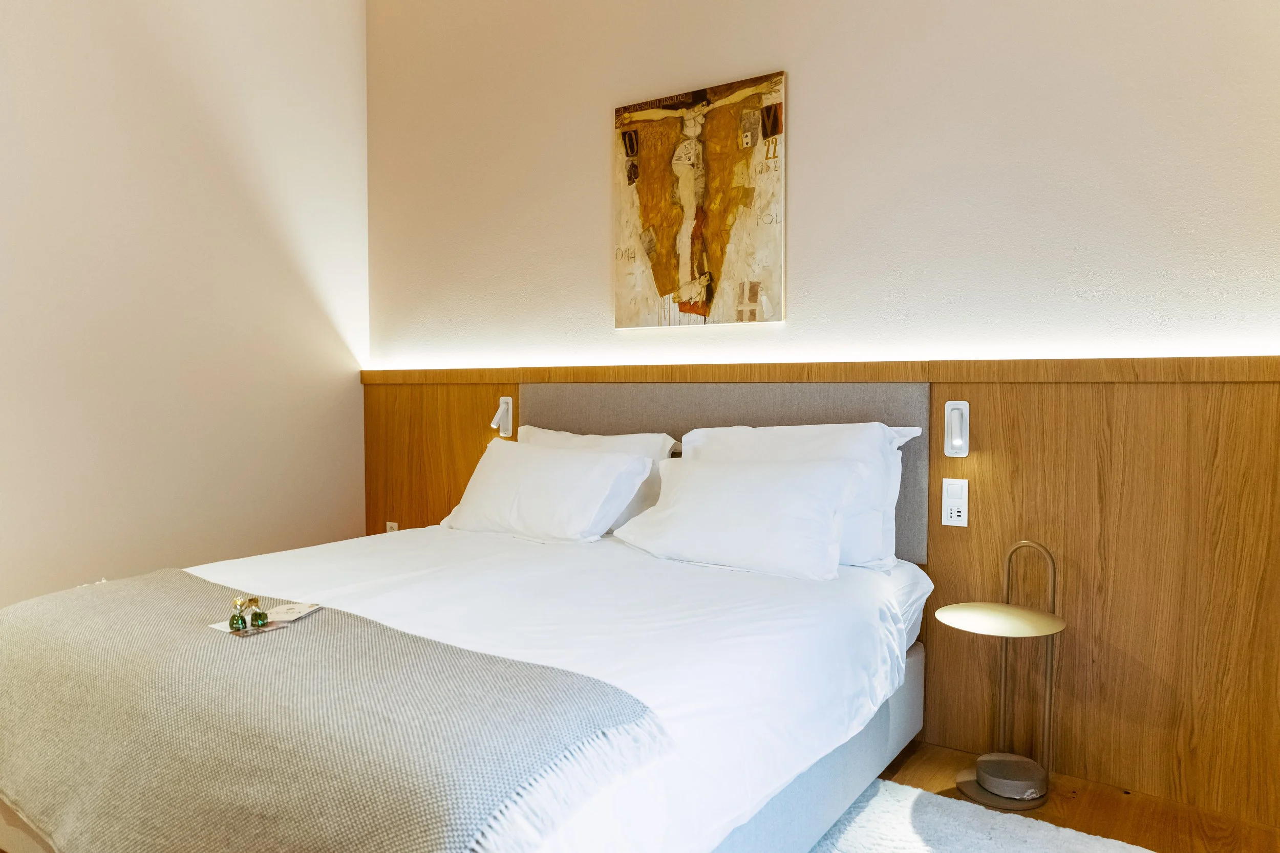 A neatly made bed with white pillows and sheets, a beige blanket, mounted reading lamps on a wooden headboard, a lighting strip above the headboard, a small side table with a gold-colored lamp, a painting above the bed, and a white rug on a wooden floor.