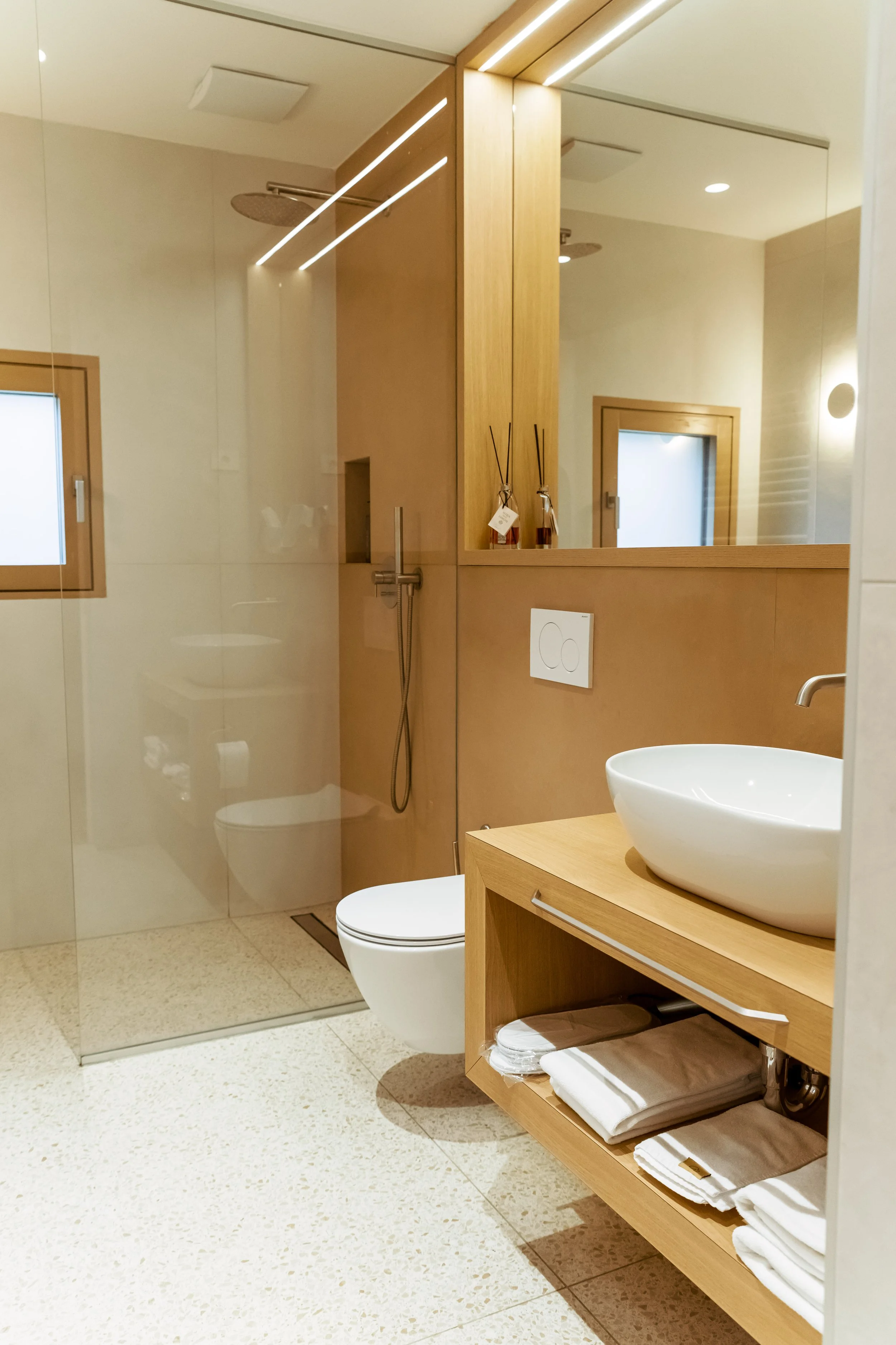 Modern bathroom with walk-in shower, wall-mounted toilet, wooden vanity with vessel sink, mirror, towels, and decorative items.