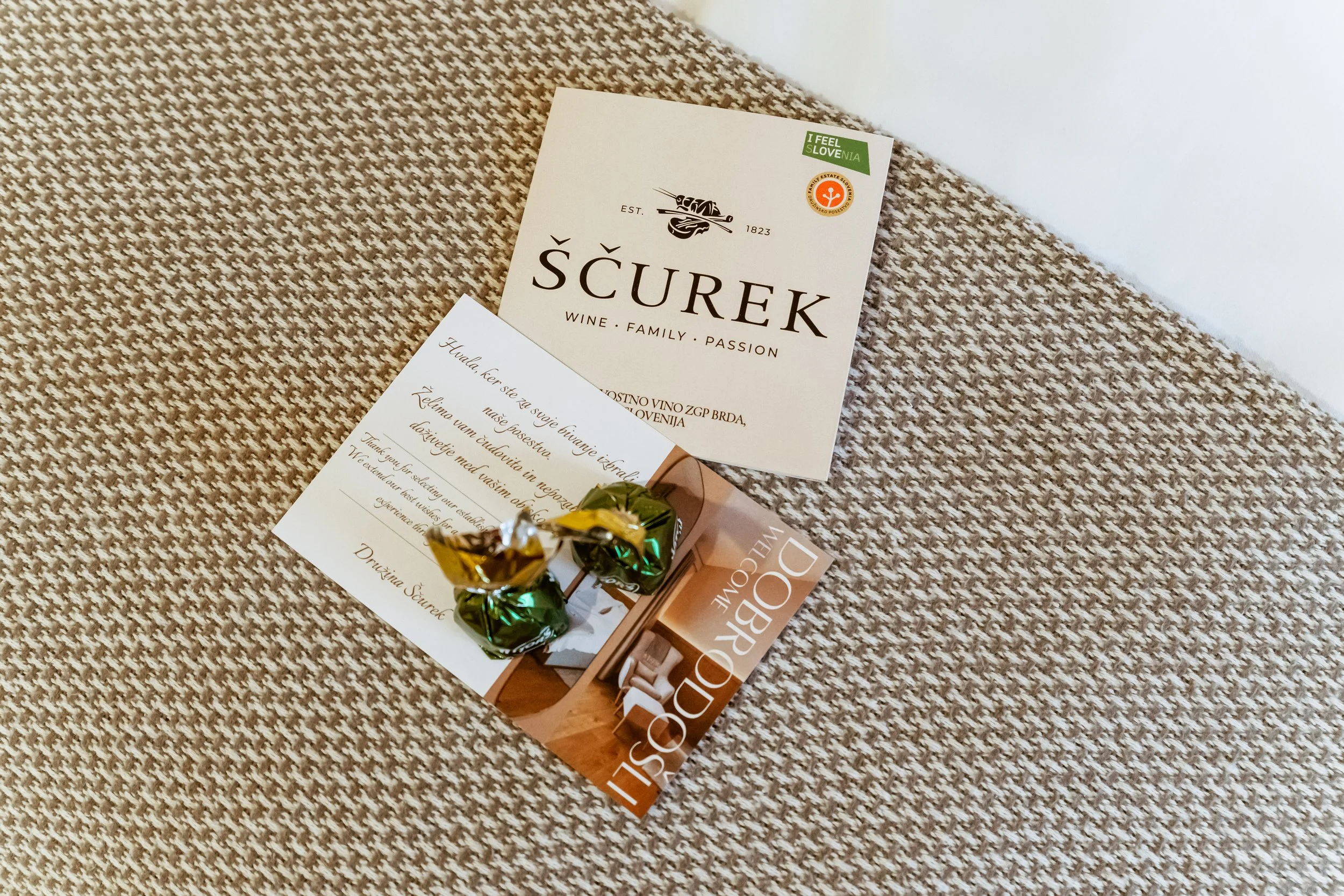 A beige textured surface with a brochure for ŠČUREK wine, a white card with Slovene text, and two small chocolates wrapped in green and gold foil.