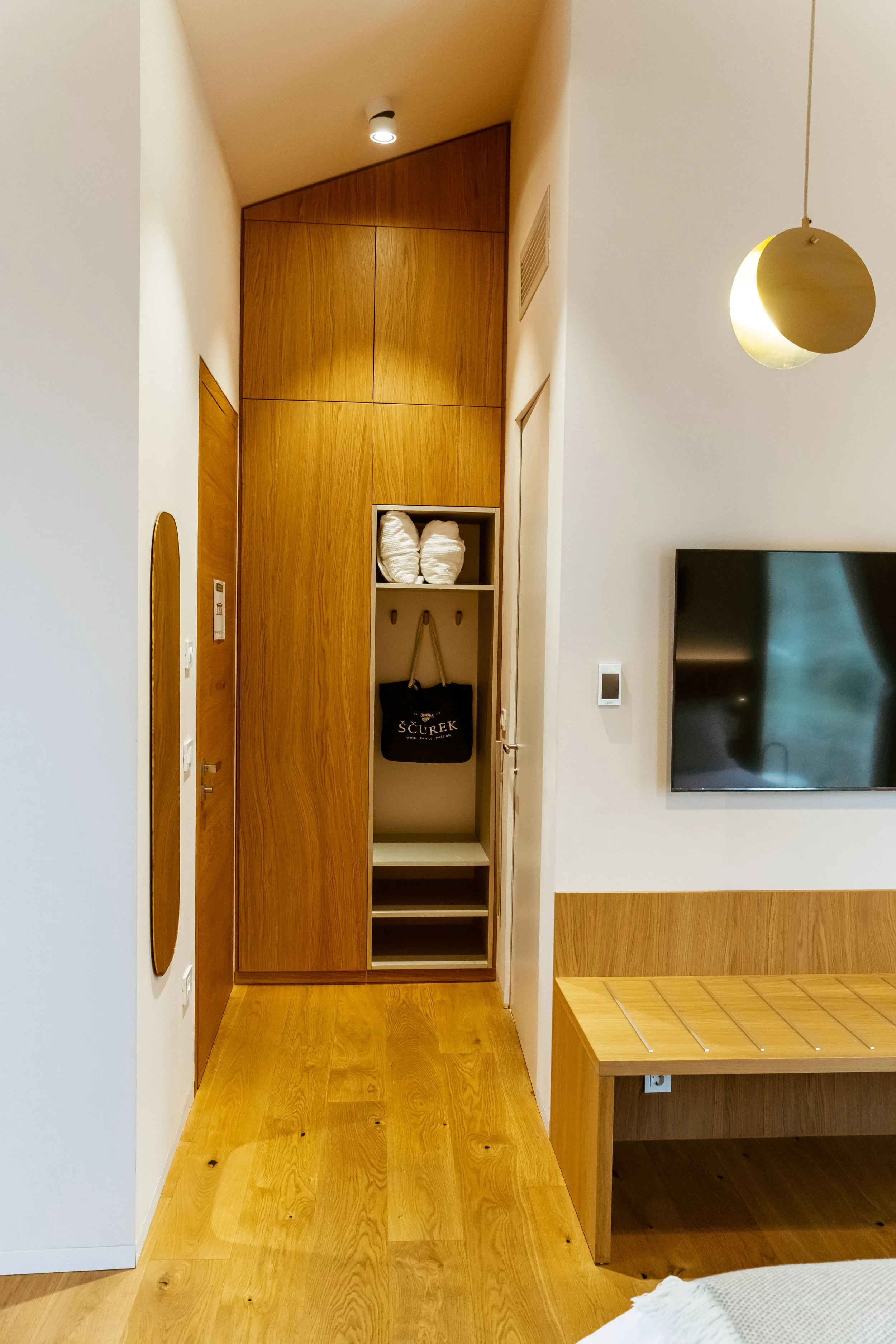 Wooden closet with shelf, towels and bag, part of a modern bedroom with white and wood furniture, wall-mounted TV, hanging pendant light, and wooden floor.