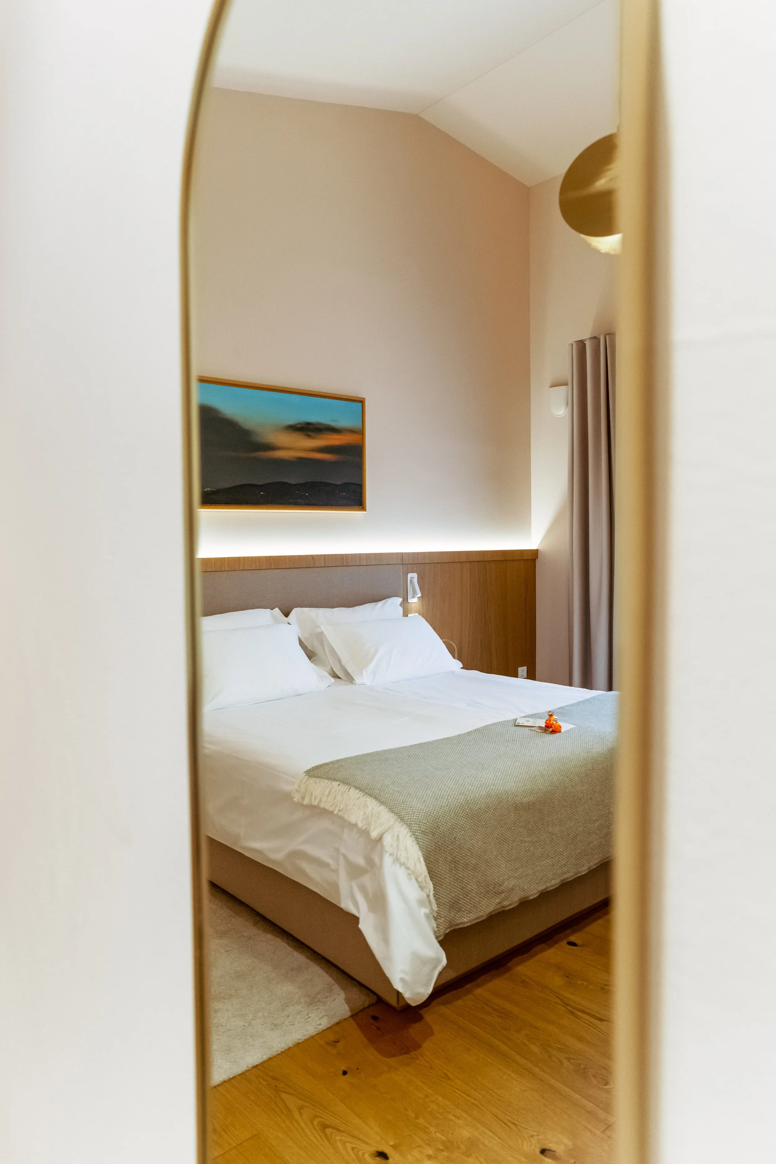 A hotel room viewed through a curved mirror showing a bed with white linens, a gray blanket, and a painting of a sunset on the wall.