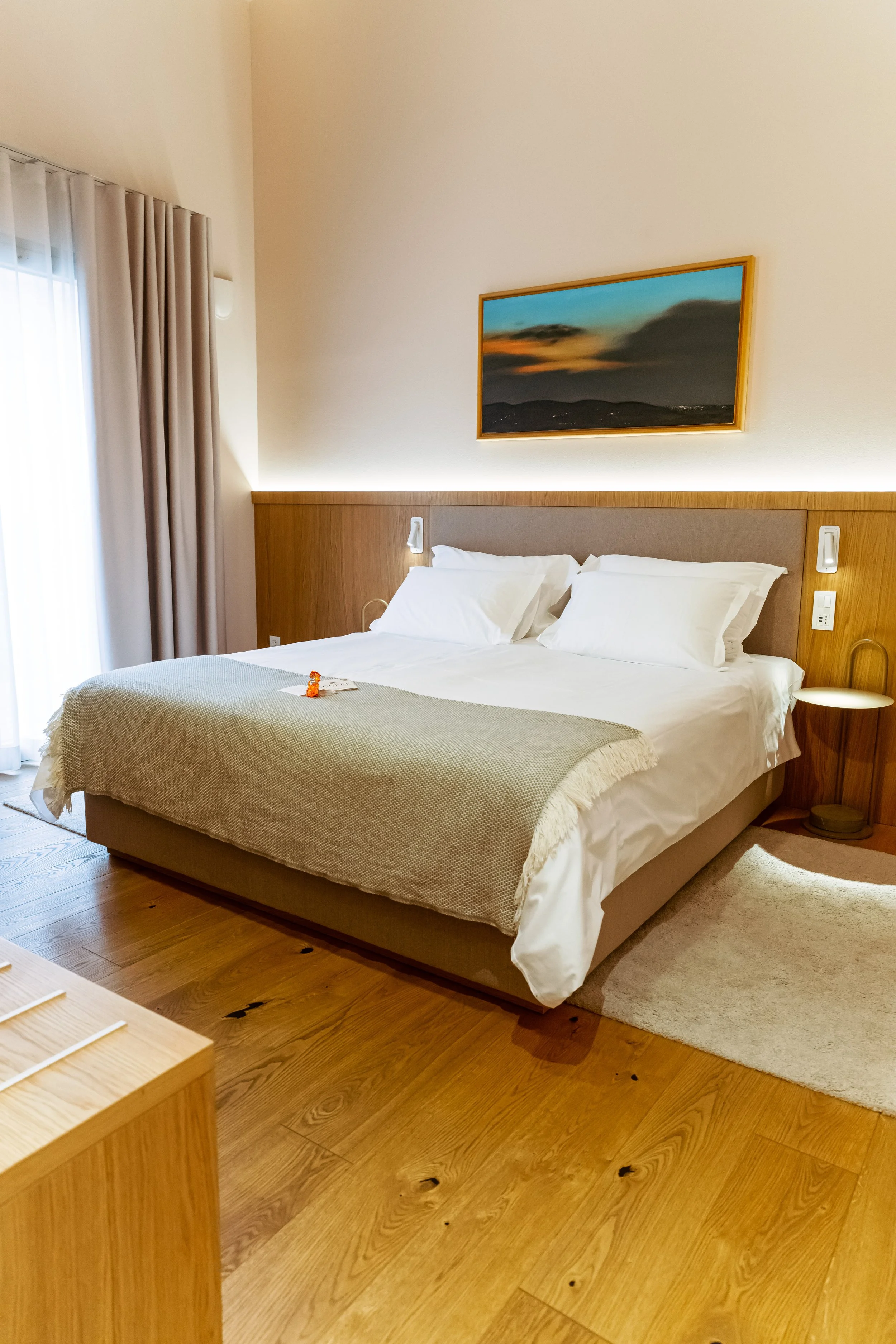 A neatly made hotel bed with white sheets, pillows, and a gray blanket in a modern hotel room with wooden flooring, a small beige rug, artwork of a sunset on the wall, and a window with curtains.