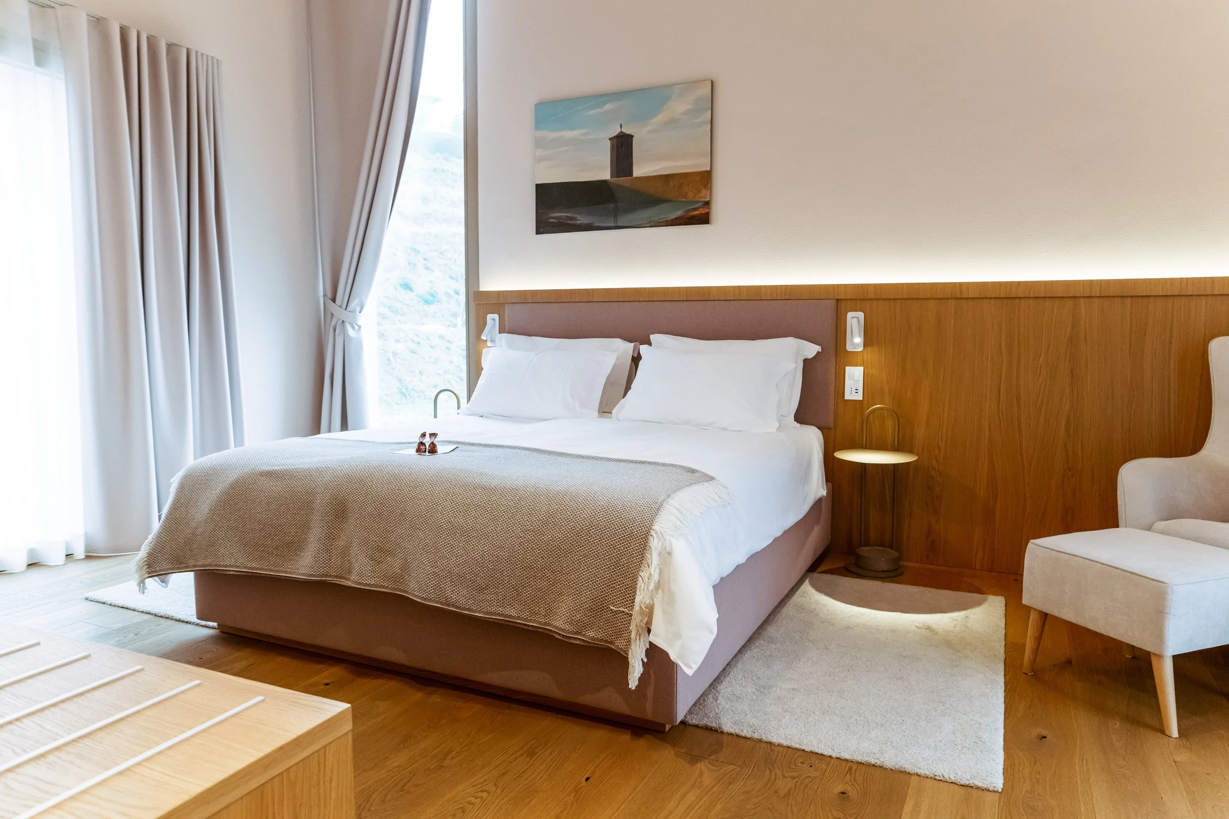 A bedroom with a large bed, white bedding, a wooden headboard, a side table with a lamp, cream curtains, a small area rug, and a piece of artwork above the bed.