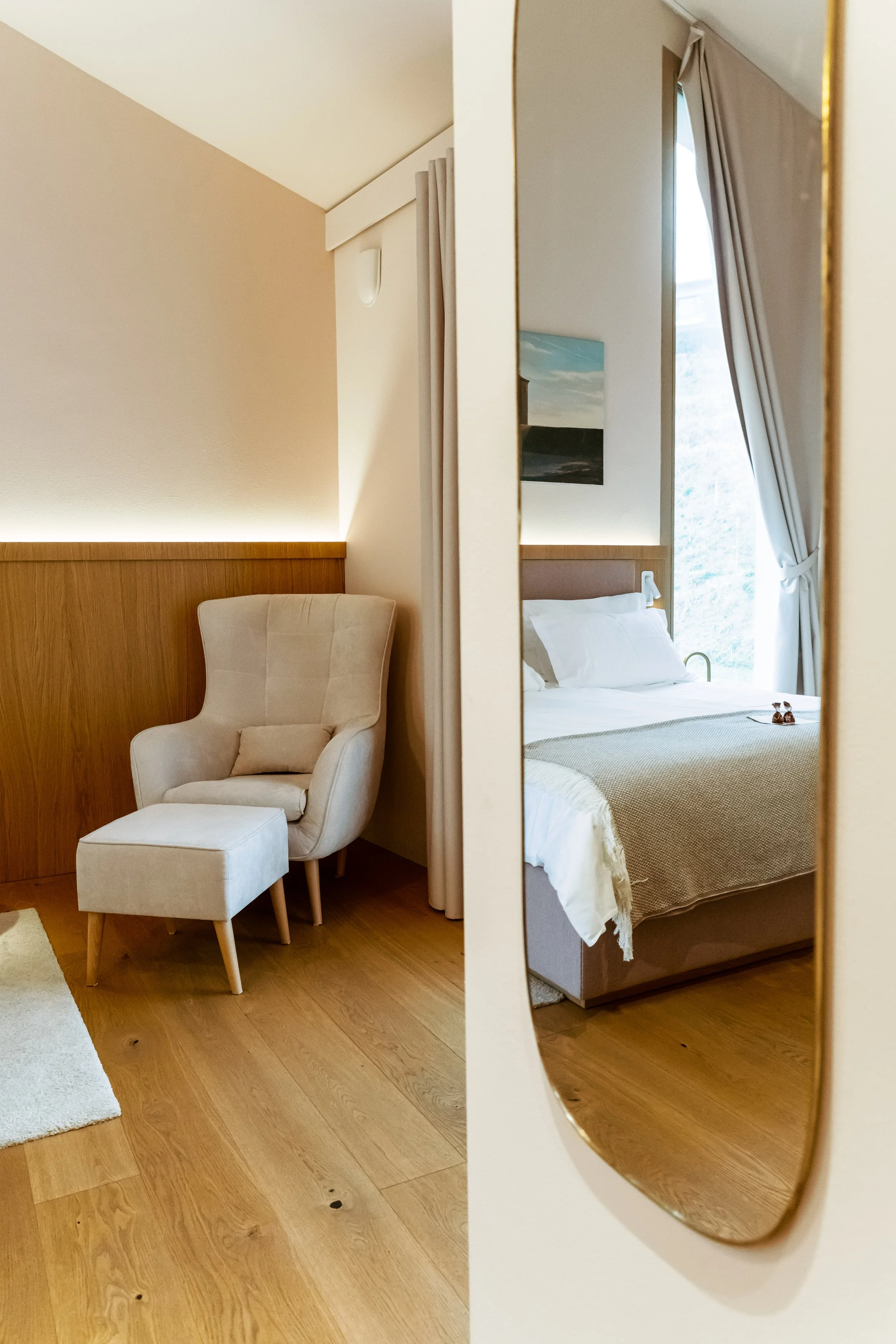 A cozy hotel room with a beige armchair and matching ottoman near a wooden wall, and a bed reflected in a full-length mirror with white linens and beige blanket, next to a window with white curtains.