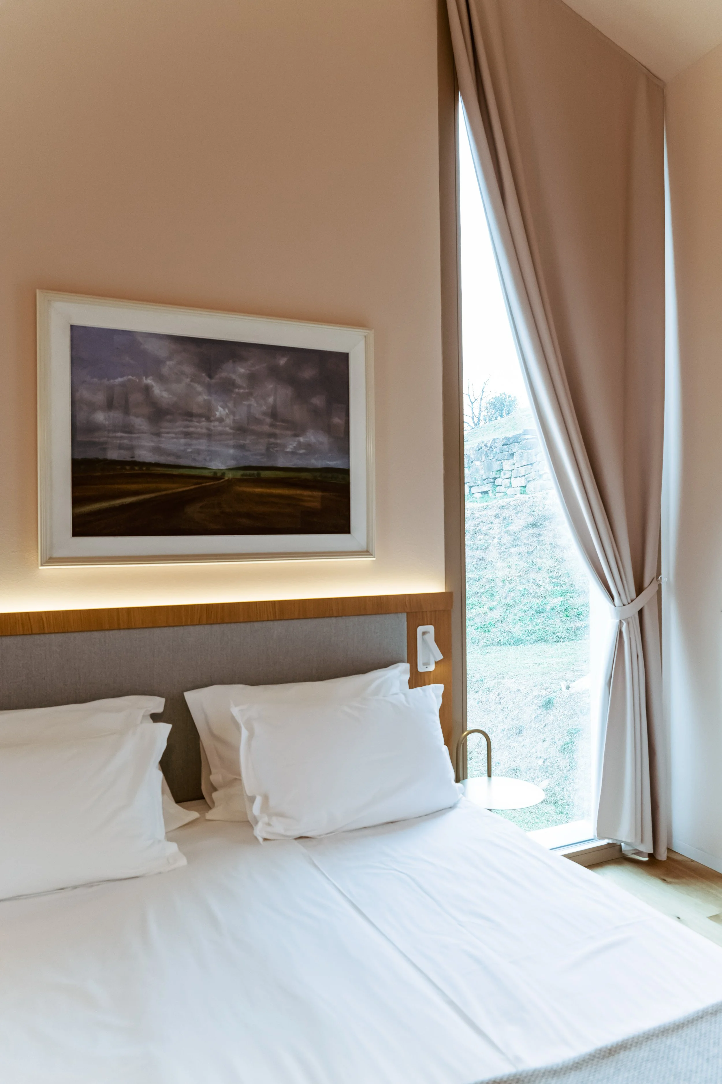 Hotel room with a bed, white pillows, a picture of a landscape with cloudy sky, and a large window with beige curtains.