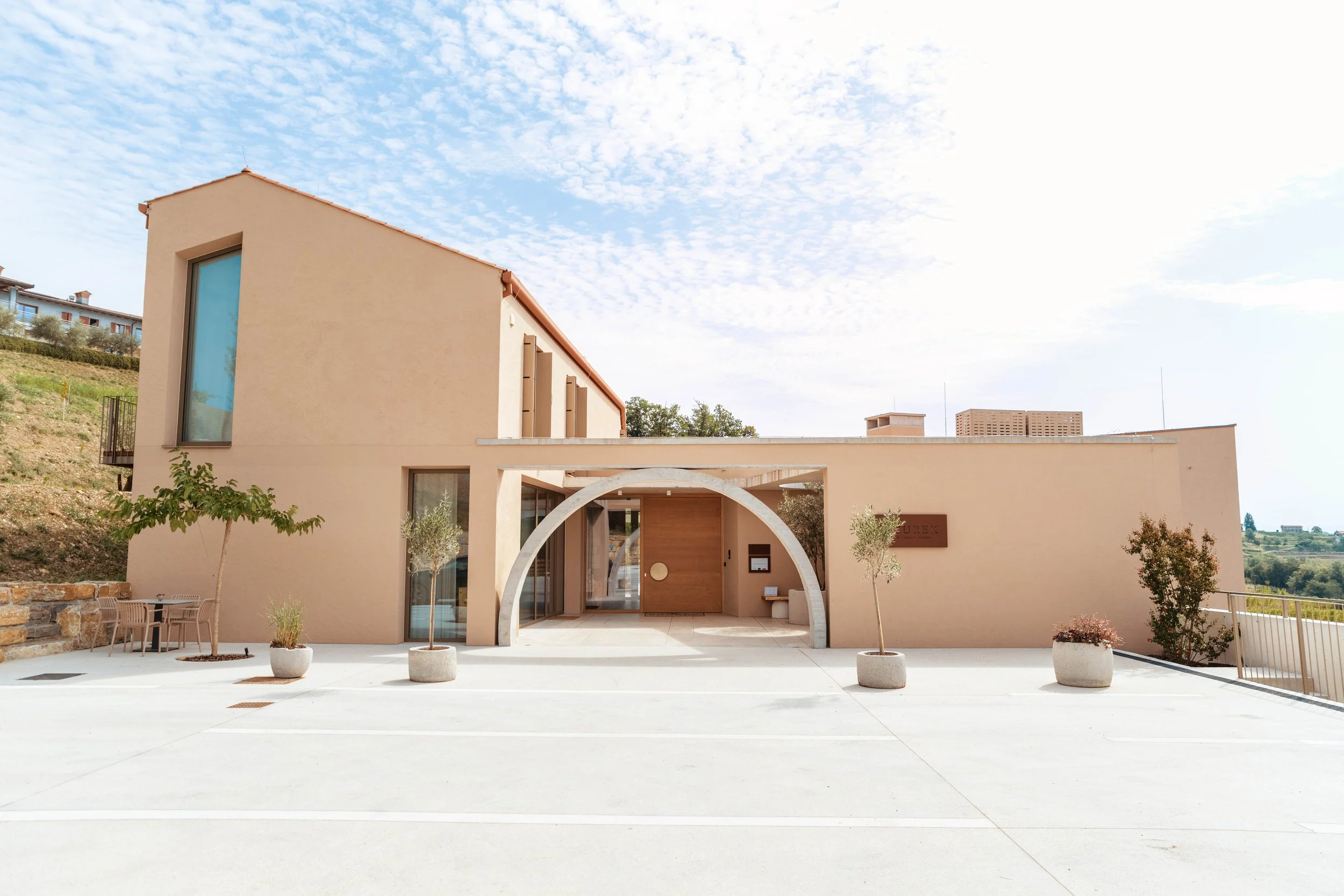 Modern beige building with an arched entrance, small trees in pots, and a parking lot in front, under a partly cloudy sky.