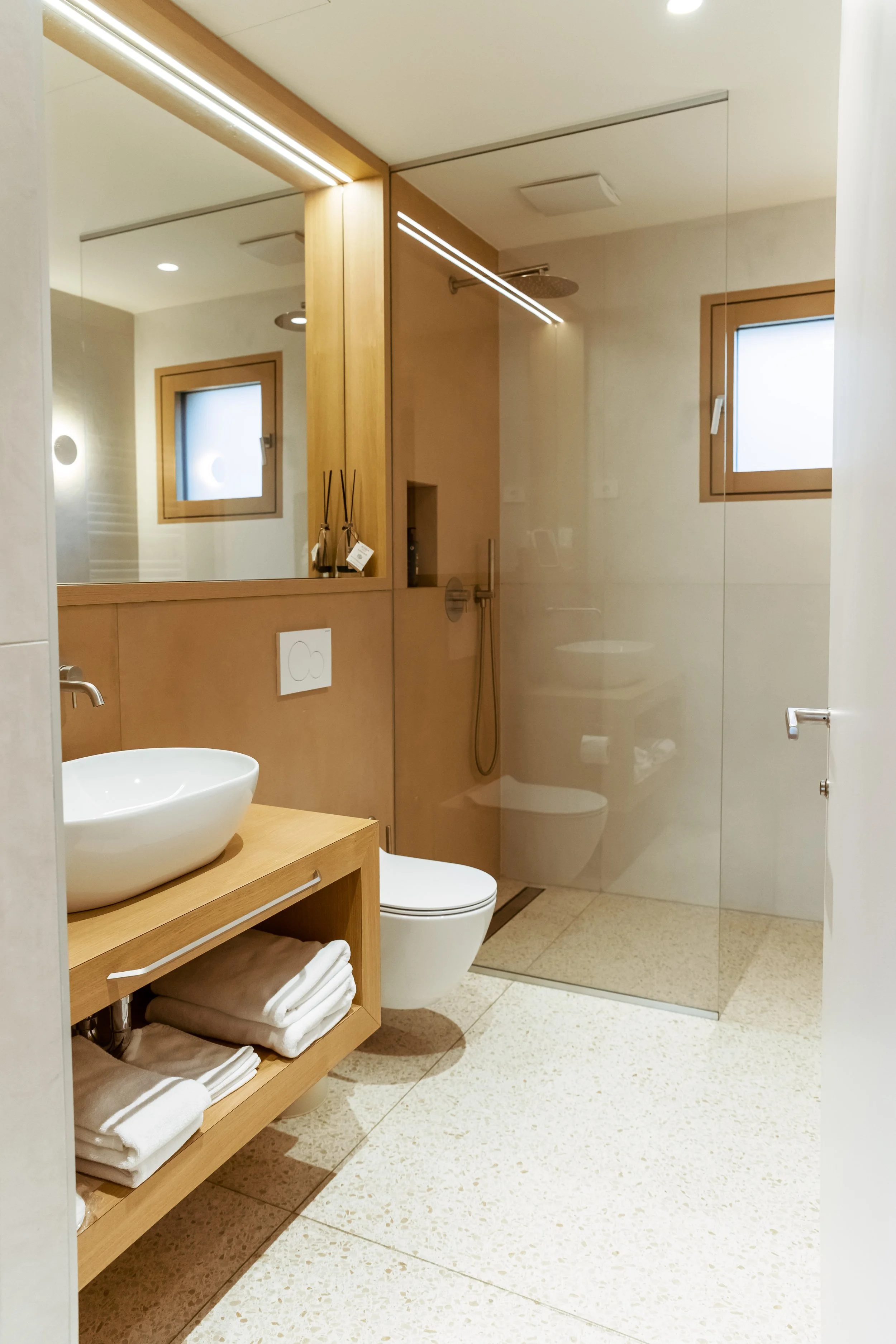 Modern bathroom with a wooden vanity, oval vessel sink, folded towels, a toilet, a glass-enclosed shower, small windows, and minimal decor.