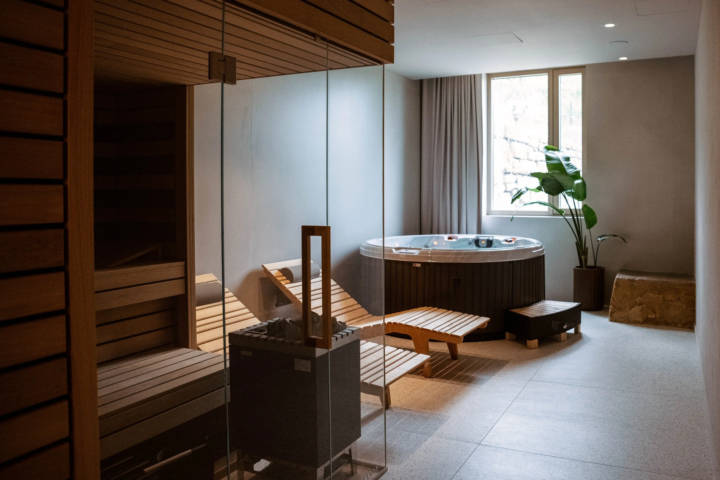 Indoor spa room with a hot tub, wooden lounge chairs, and a sauna. Large window with curtains, and a potted plant near the window.