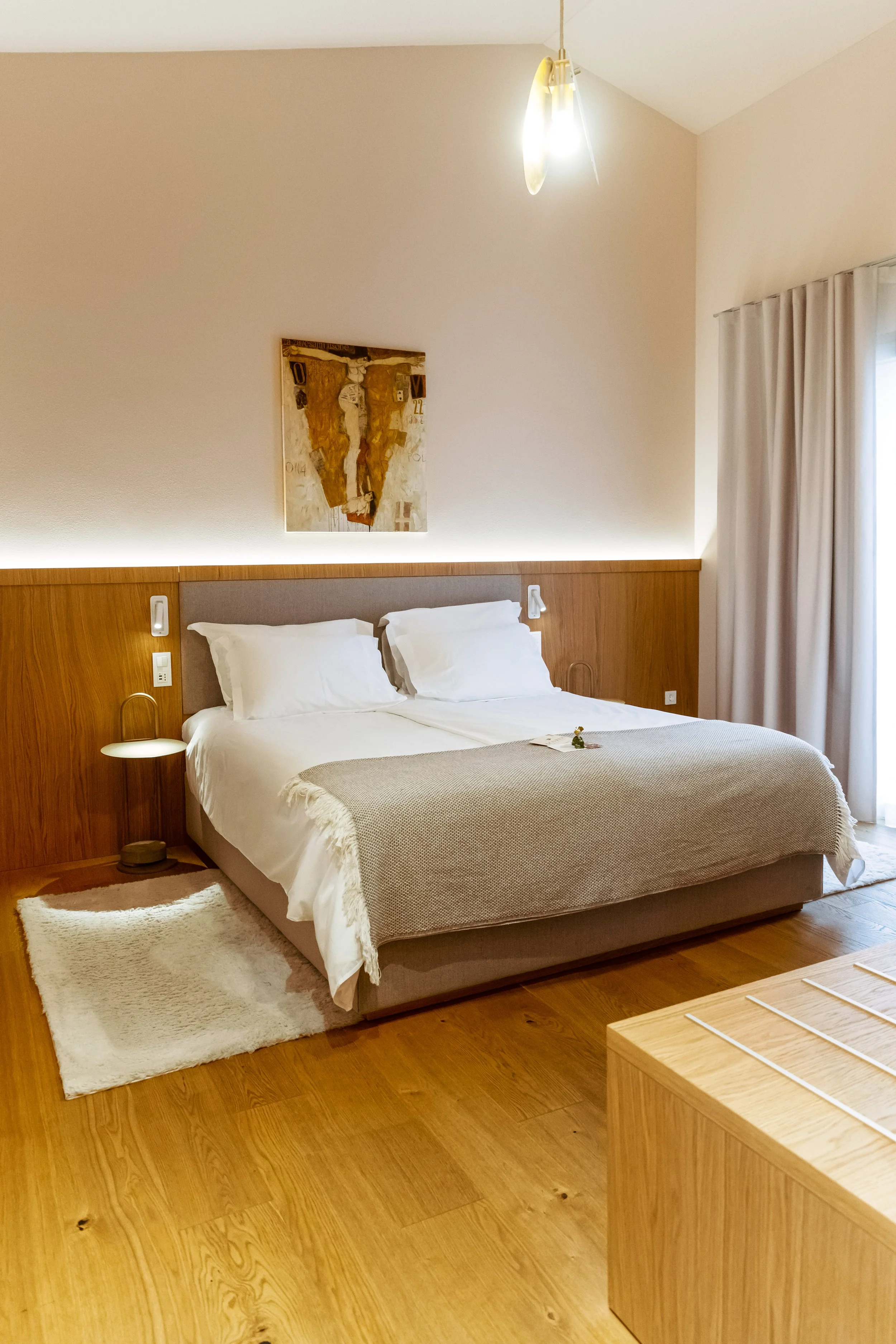 A neatly made hotel bedroom with a large bed featuring white bedding and a beige blanket, a small bedside table with a lamp, framed artwork above the bed, and a set of beige curtains on the window, all with wooden flooring.