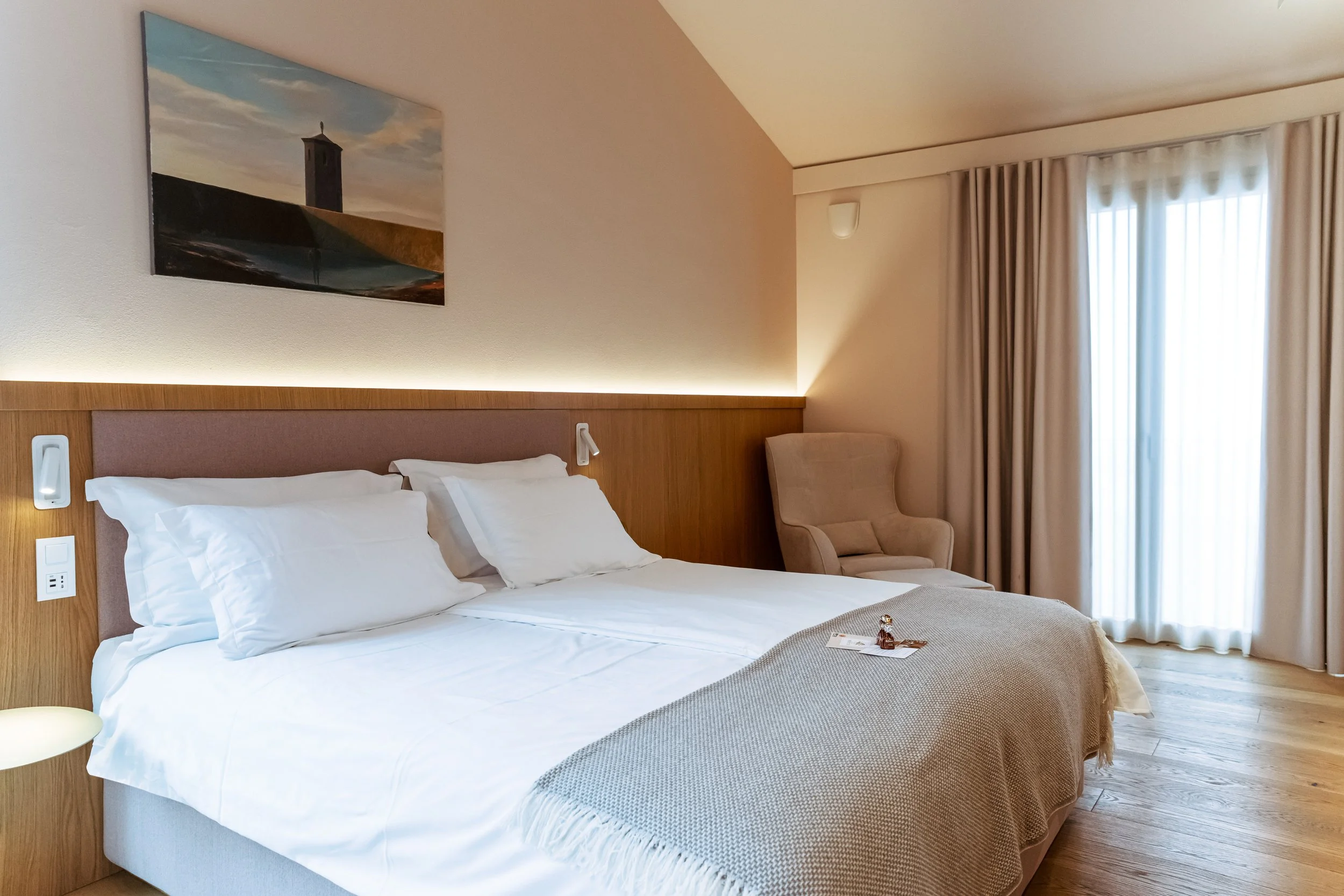 A hotel room with a bed, beige walls, a painting above the headboard, a large window with curtains, and a beige armchair in the corner.