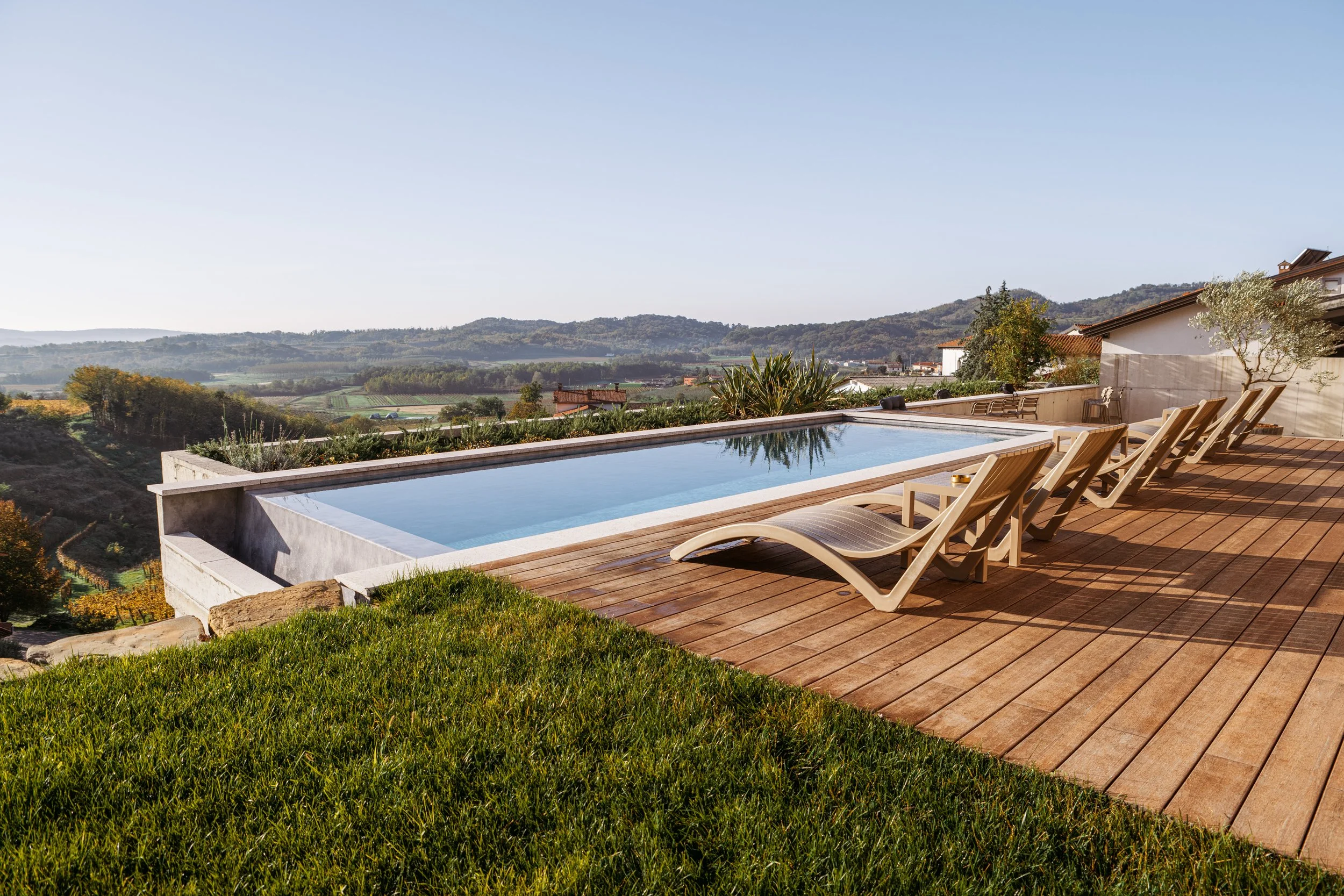 Luxury outdoor swimming pool with lounge chairs on a wooden deck overlooking a scenic countryside landscape with rolling hills, houses, and trees.