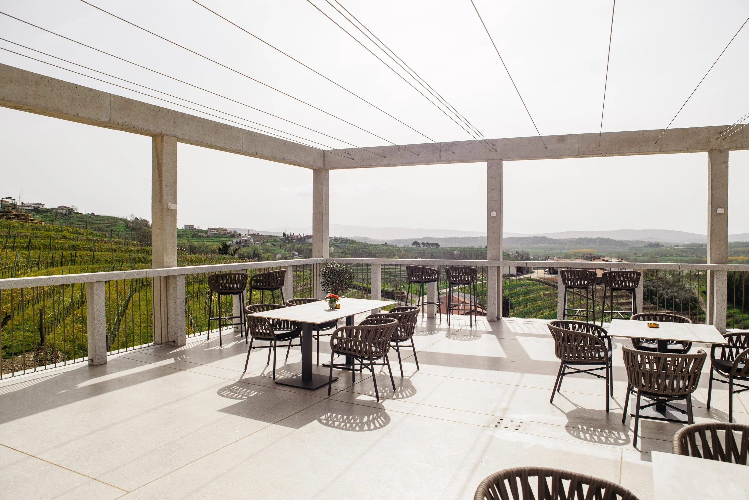 Empty outdoor patio with white tables and black chairs overlooking a green landscape with hills and vineyards.