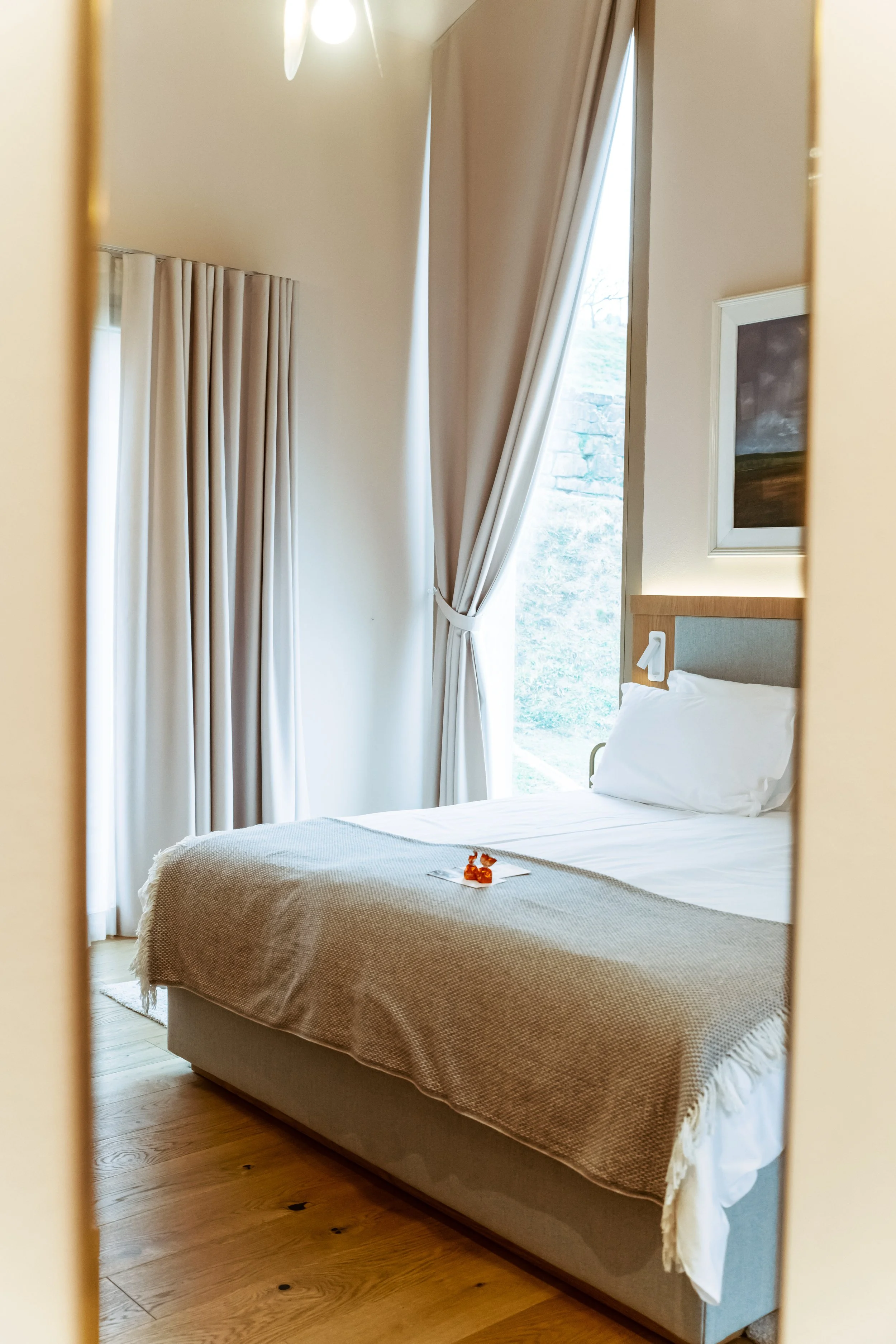 A neatly made hotel bed with white sheets and a beige blanket, reflected in a mirror, with curtains and a window in the background.