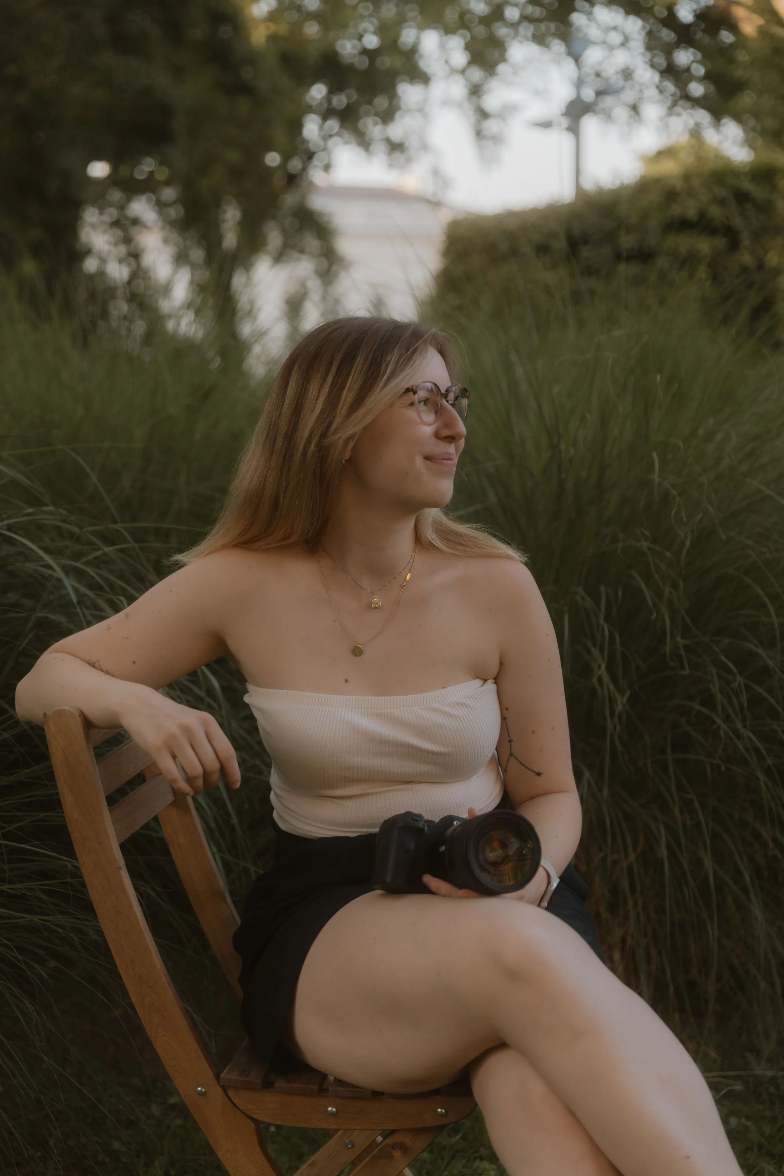 A woman with long blonde hair and glasses sitting outdoors on a wooden chair, holding a camera, with greenery and trees in the background.