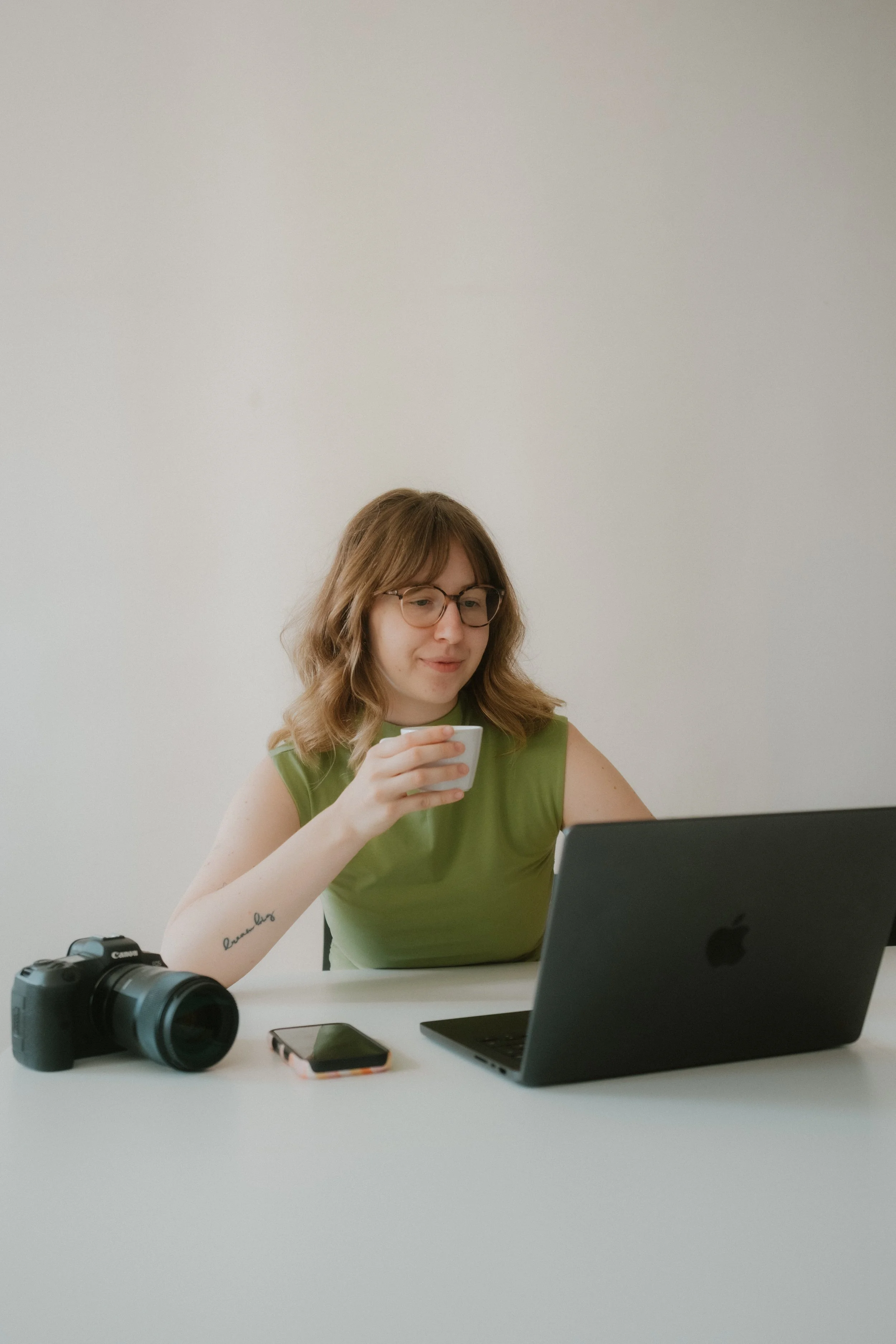 Woman in eyeglasses sitting at white table with a laptop, camera, smartphone, and coffee cup, working on her computer.