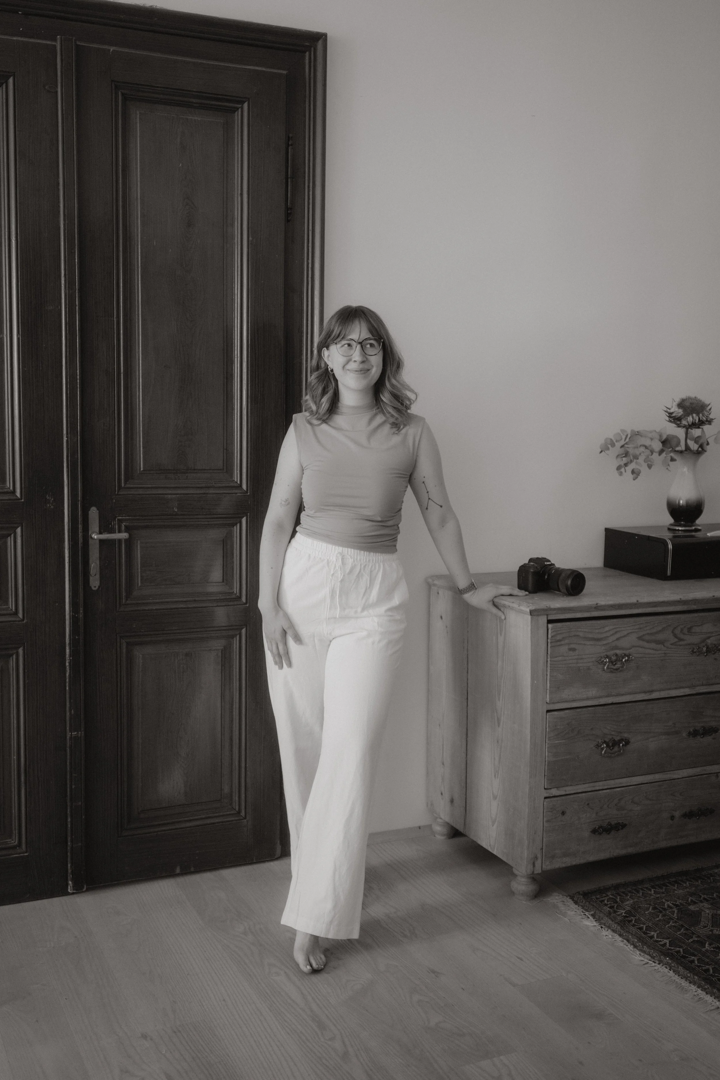 A woman with glasses and wavy hair stands indoors, smiling and leaning against a wooden dresser with a camera on top. There is a dark wooden door to her left and a vase with flowers on the dresser.