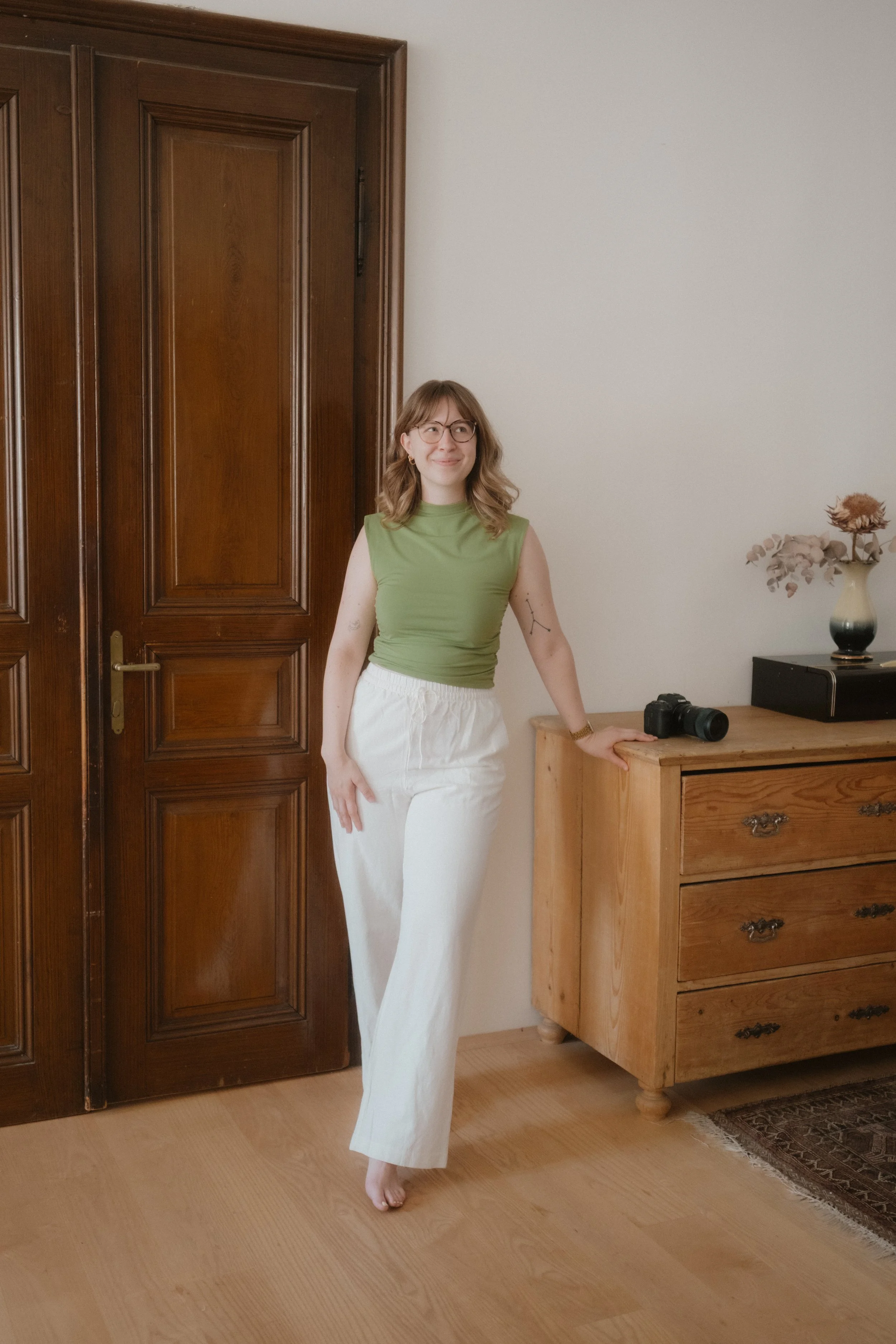 A woman with shoulder-length wavy hair, glasses, wearing a green sleeveless top and white pants, standing in a room next to a wooden dresser, with a camera on top of the dresser and a vase with flowers on it, and a wooden door in the background.