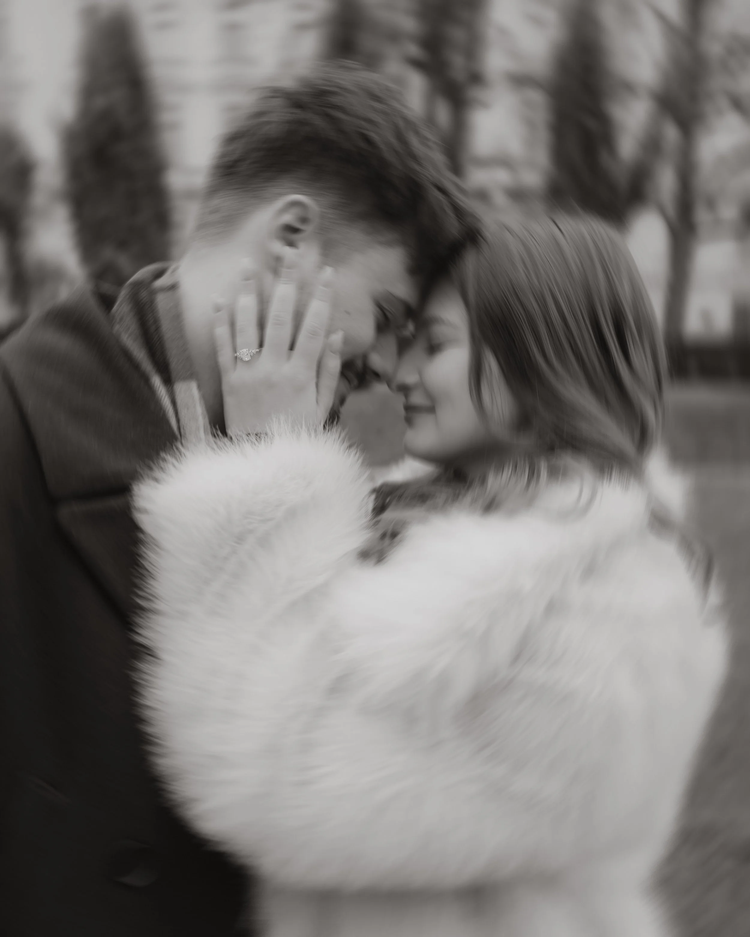 Close-up of a man and woman smiling with foreheads touching, outdoors with blurred trees in background.