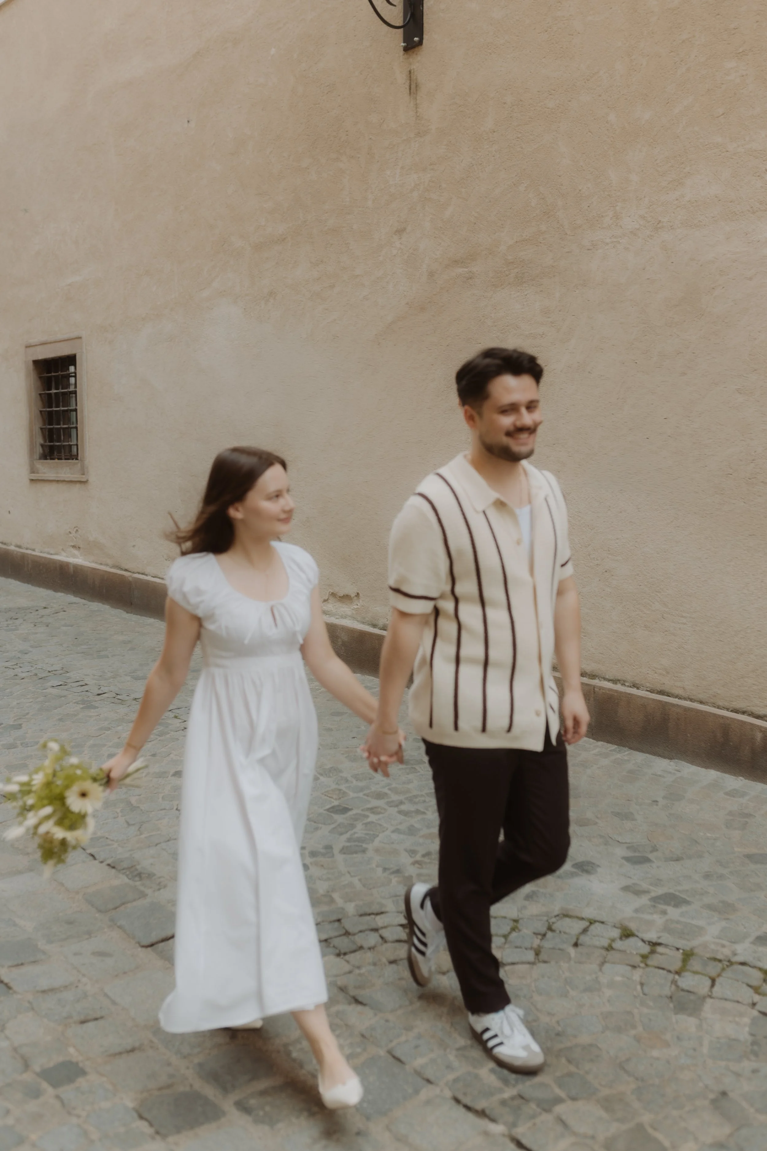 A couple walking hand in hand on a cobblestone street, the woman wearing a white dress and holding flowers, and the man wearing a beige striped shirt and black pants, smiling and looking content.