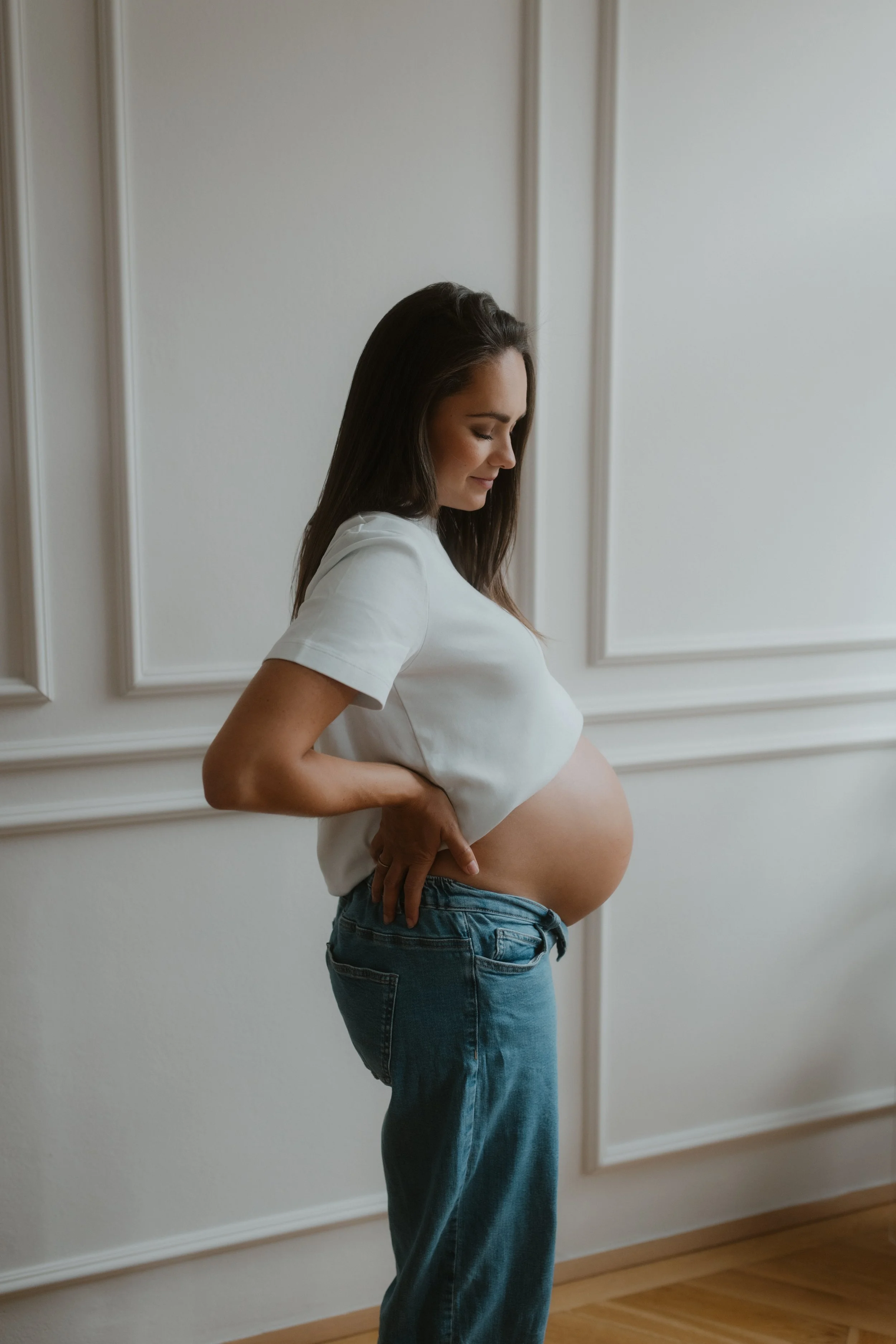 A pregnant woman with long brown hair wearing a white t-shirt and blue jeans is standing indoors against a white wall with decorative molding, smiling gently and looking down at her belly.