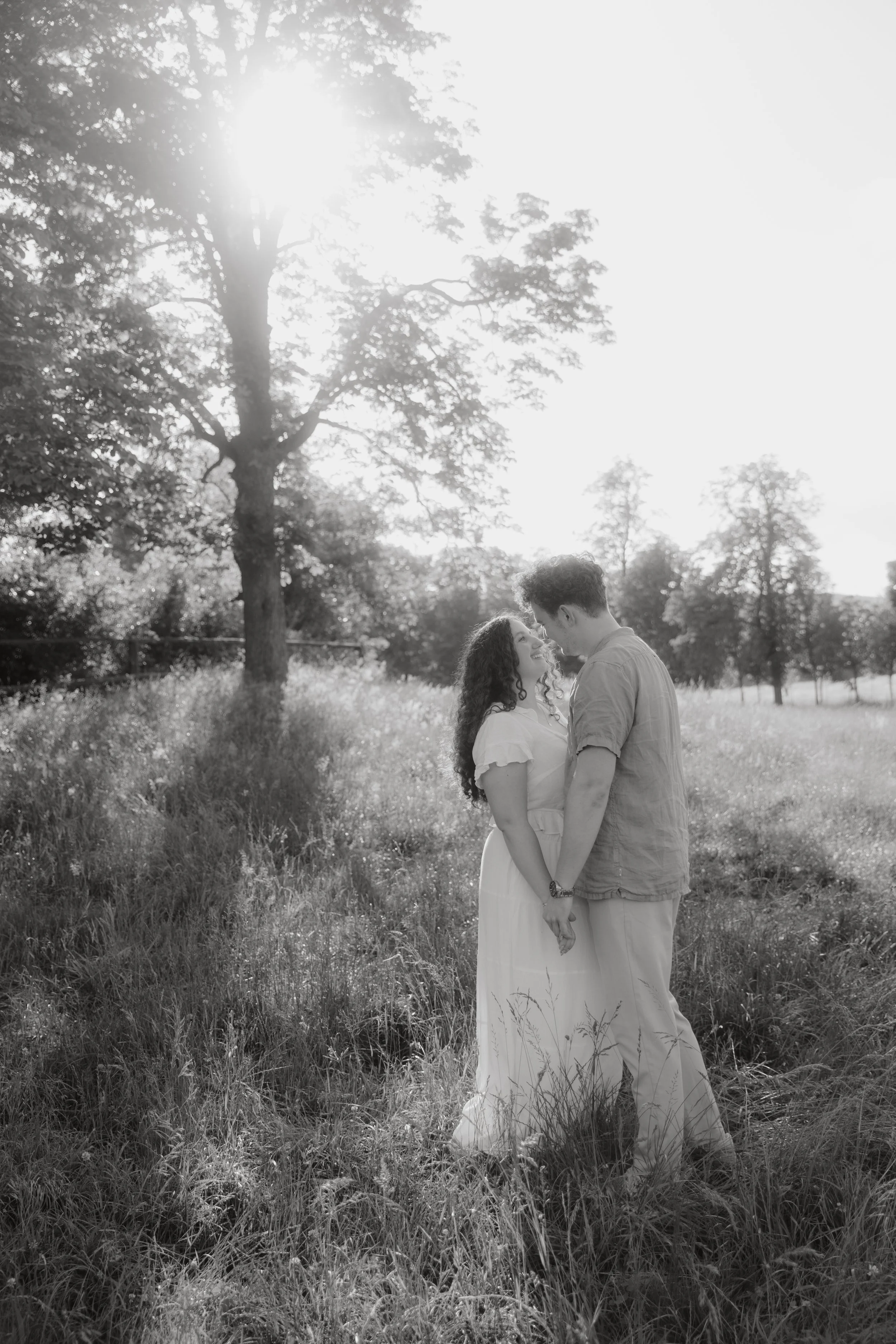 A couple standing close together in a grassy field with trees in the background, captured in black and white with sunlight filtering through the trees.