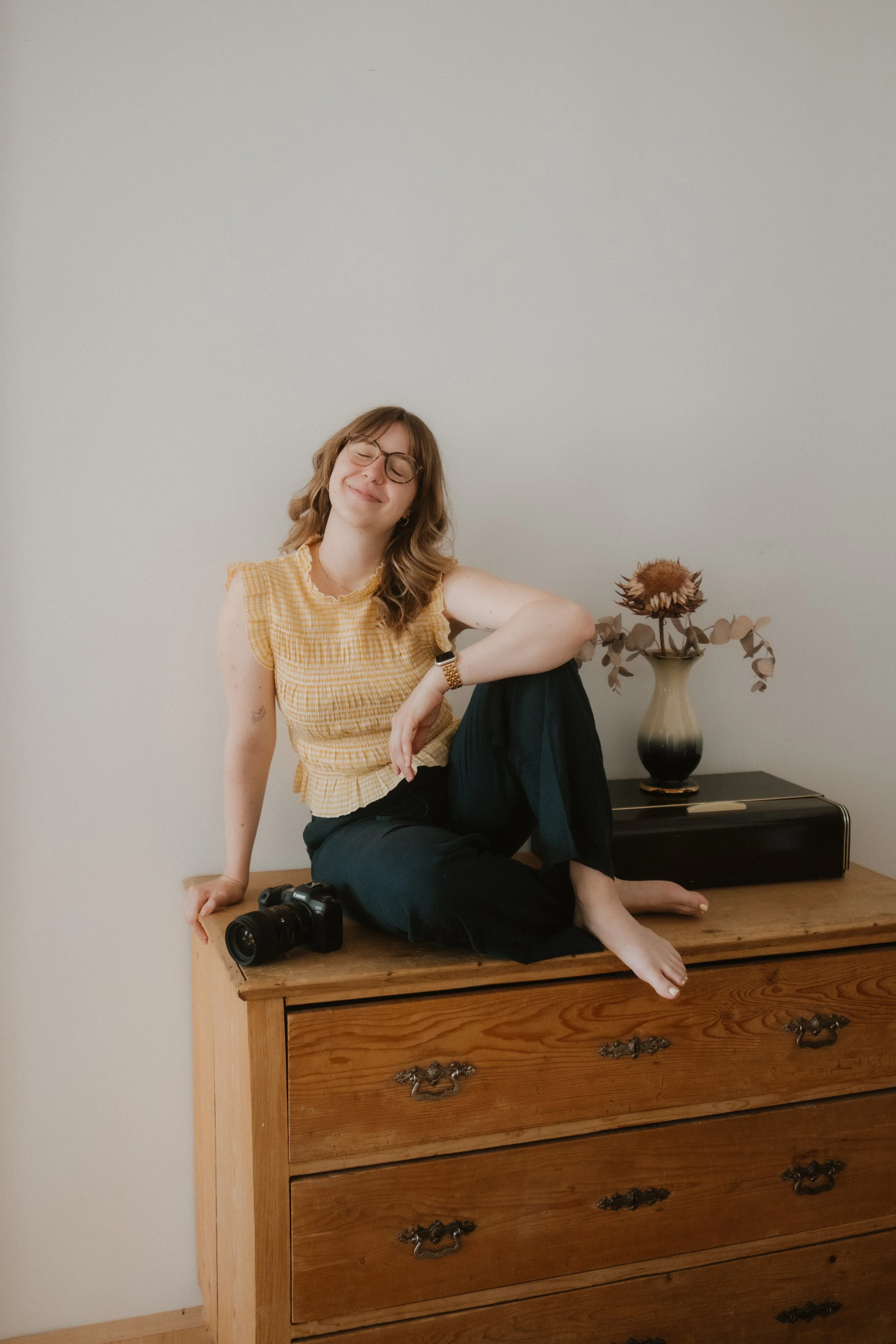 A young woman with glasses, brown hair, and a yellow checkered top sitting on a wooden dresser, smiling with eyes closed. She has a camera resting on the dresser and is sitting next to a black and beige vase with dried flowers on top. The background is a plain light-colored wall.