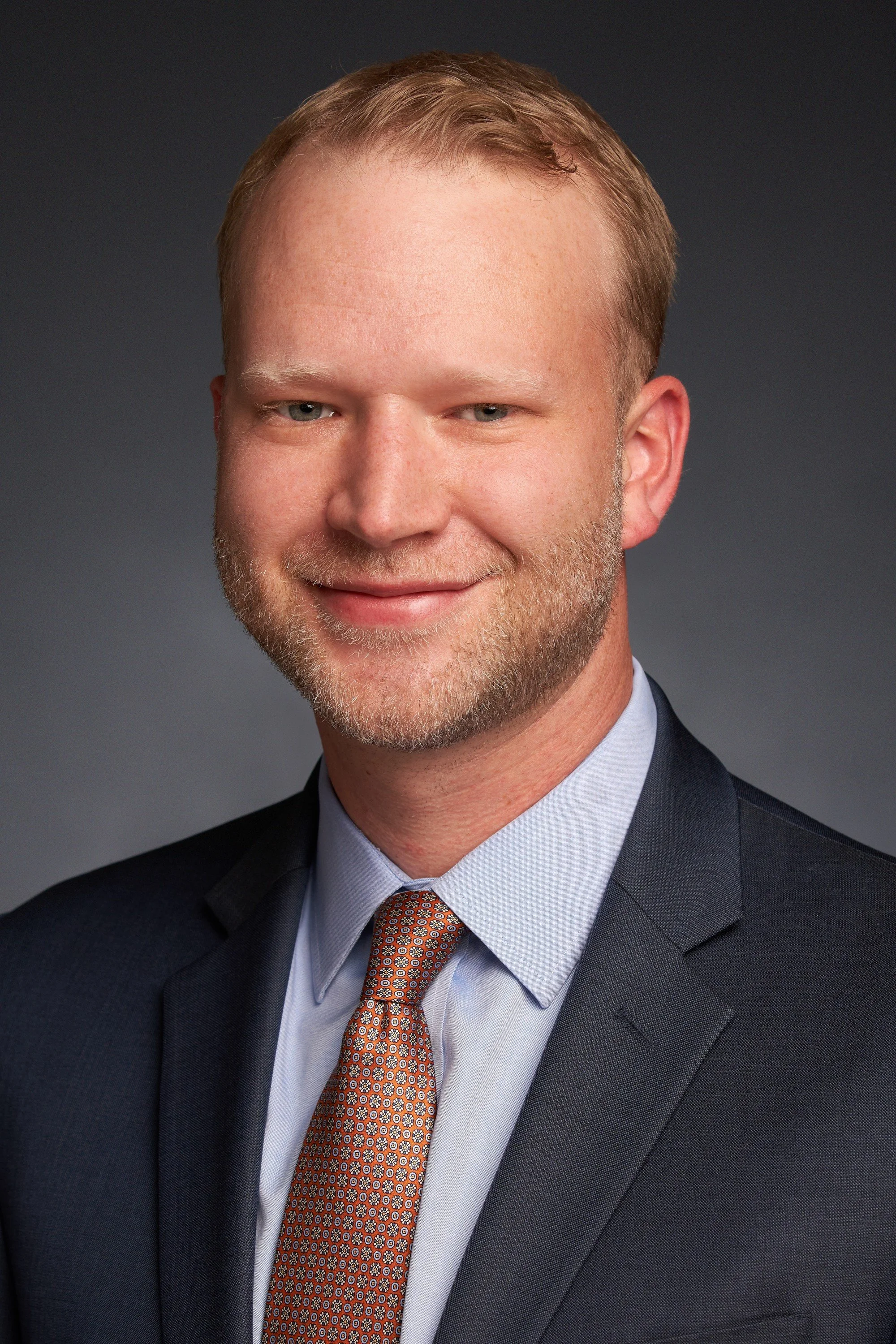 An elderly man in religious attire, possibly a priest or minister, standing indoors, wearing a white robe with a cincture. He is smiling and part of a group of people in what appears to be a church setting with stained glass windows.