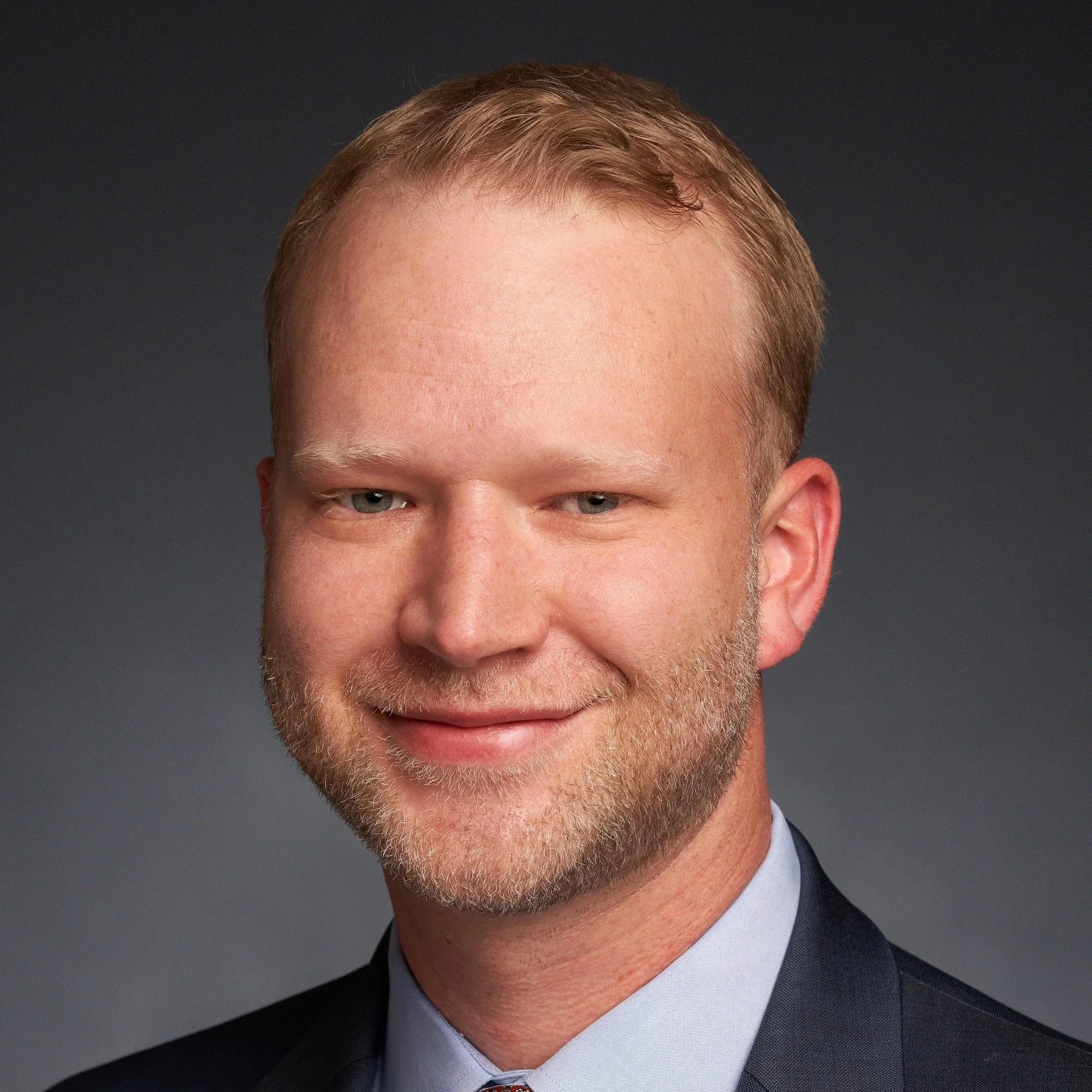 An elderly man in religious attire, possibly a priest or minister, standing indoors, wearing a white robe with a cincture. He is smiling and part of a group of people in what appears to be a church setting with stained glass windows.