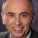 Businessman wearing glasses and a suit with a blue striped tie, posing indoors with a blurred background.