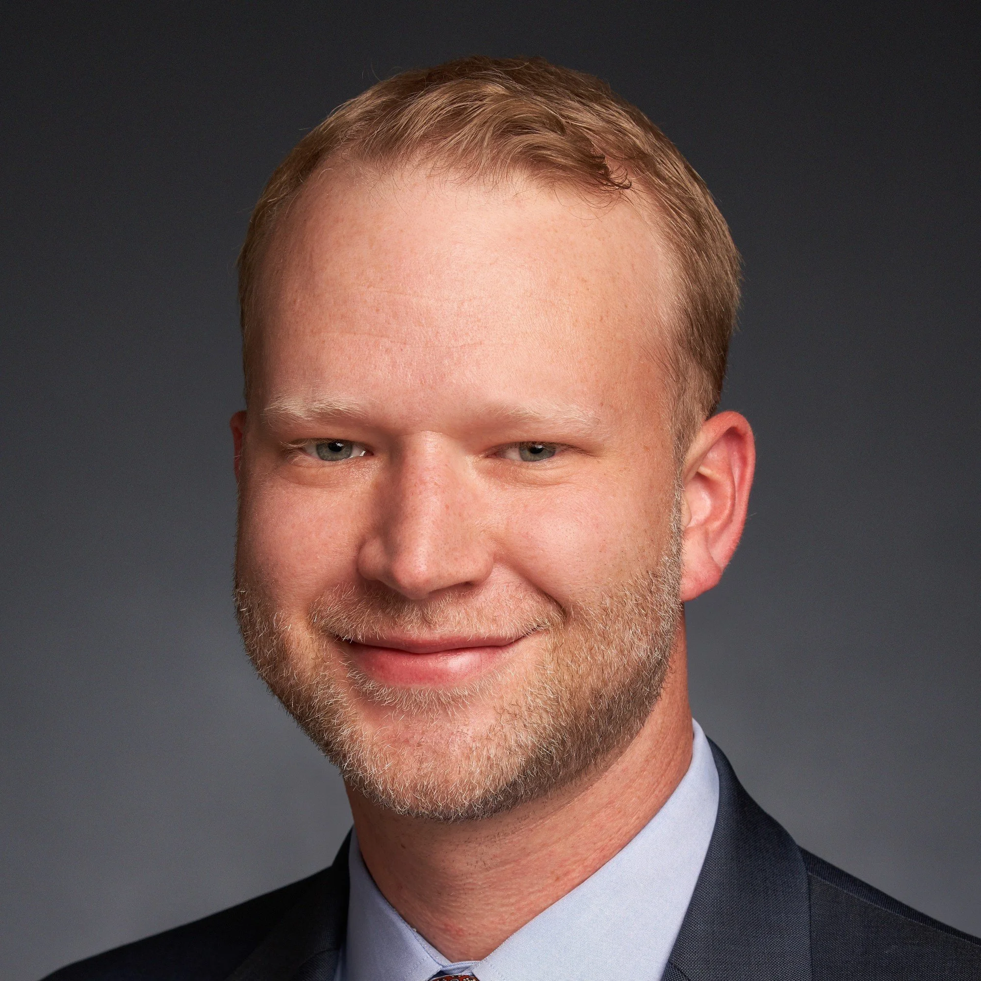 An elderly man in religious attire, possibly a priest or minister, standing indoors, wearing a white robe with a cincture. He is smiling and part of a group of people in what appears to be a church setting with stained glass windows.
