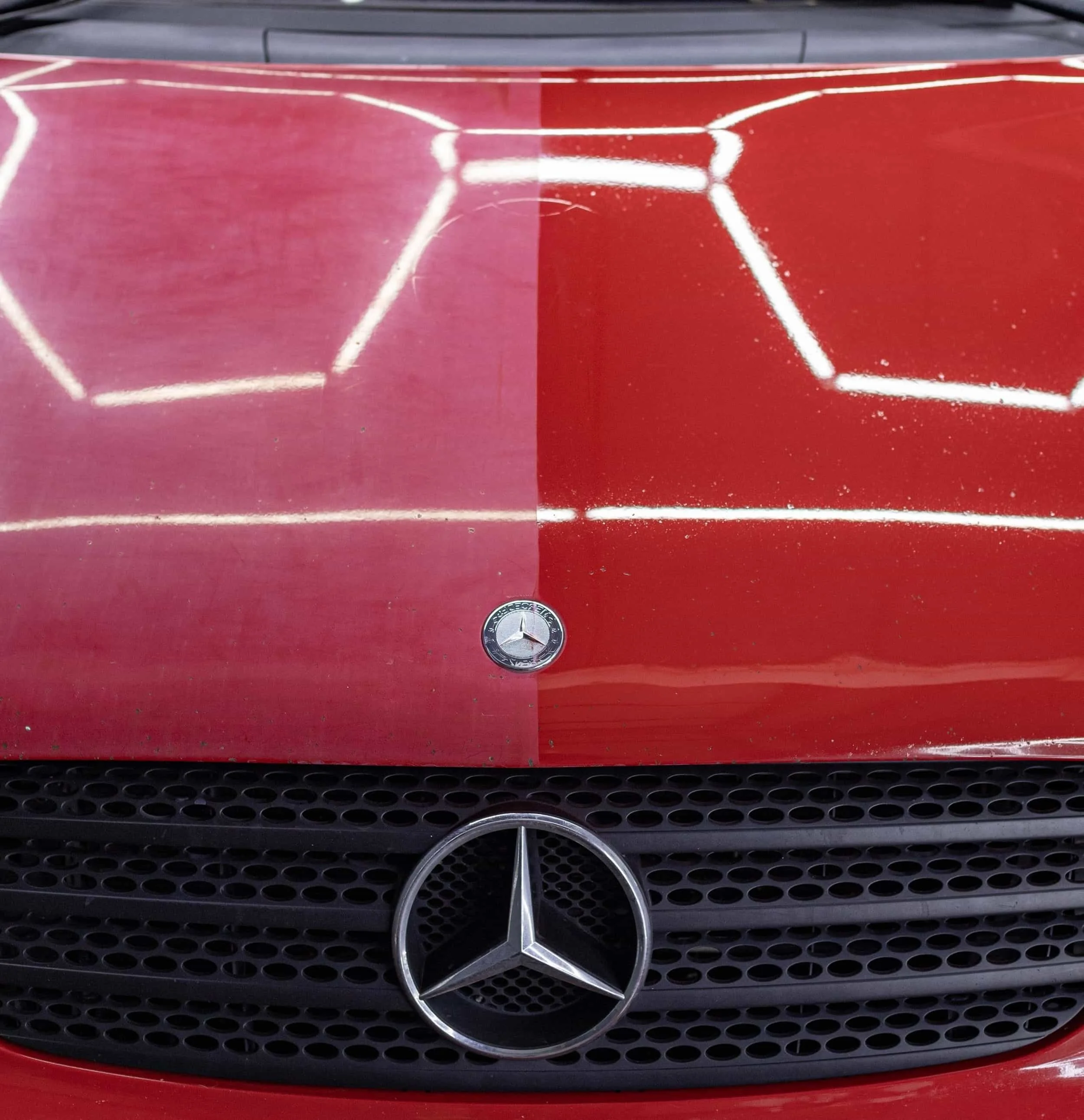 Front of a red Mercedes-Benz car with a Mercedes-Benz emblem on the hood and grille, showing a split reflection of the hood with one half polished and the other undone