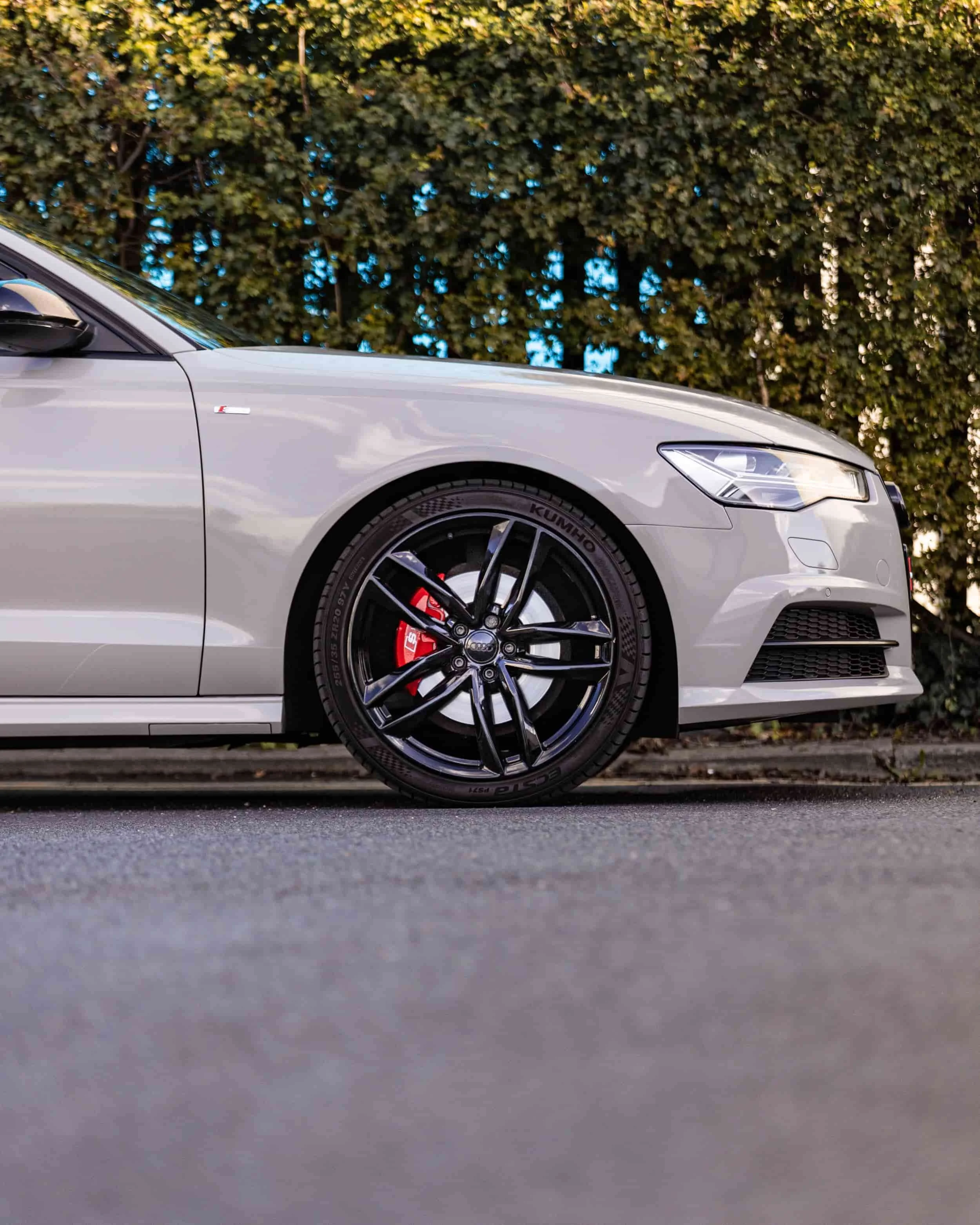 Close-up of a silver sports car's front left side, showing a black alloy wheel with a red brake caliper behind it, and a background of green trees.