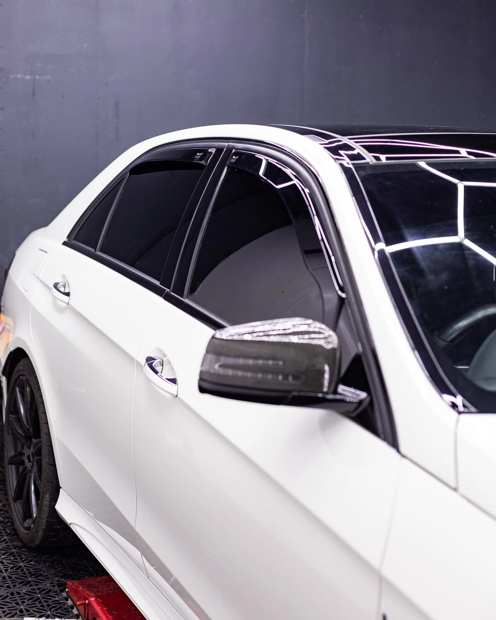 White luxury car with tinted windows is parked indoors under bright geometric ceiling lights.