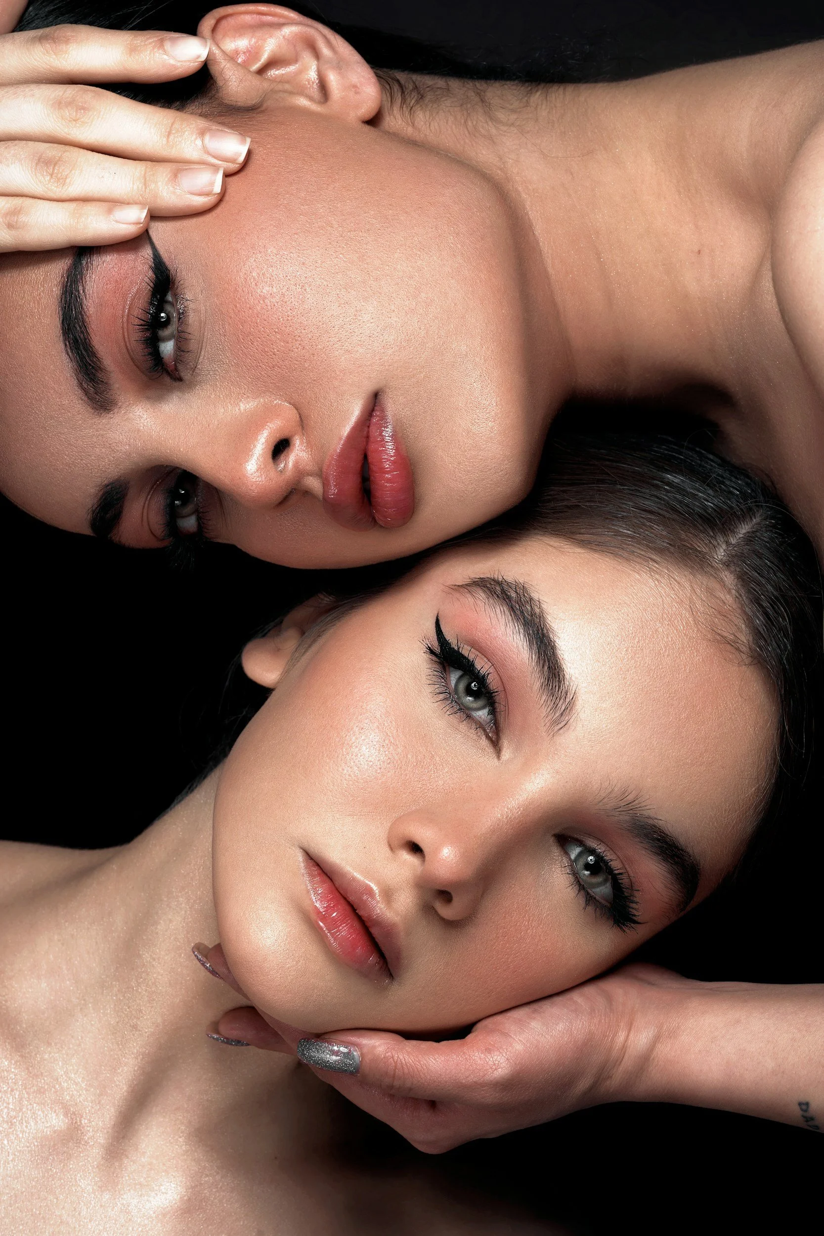 Two women with makeup, one with her hand on her forehead and the other resting her face on her hand against a black background.