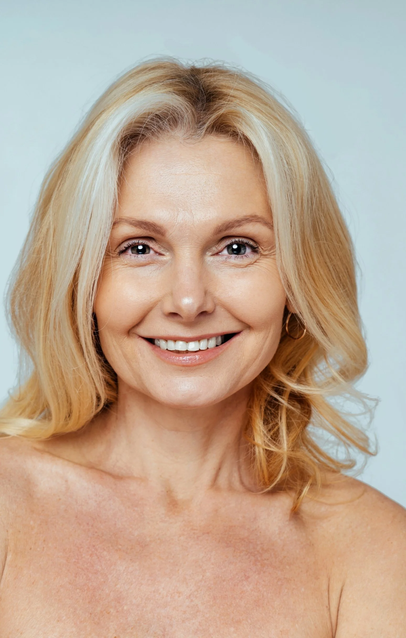Close-up of a smiling woman with blonde hair, blue eyes, and light skin, showing her bare shoulders.