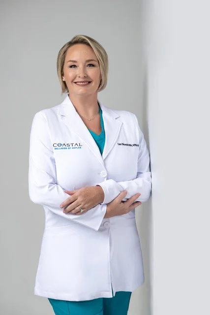 A smiling woman wearing a white medical coat with the logo 'Coastal Wellness of Naples' and teal scrubs underneath, standing indoors against a plain wall.
