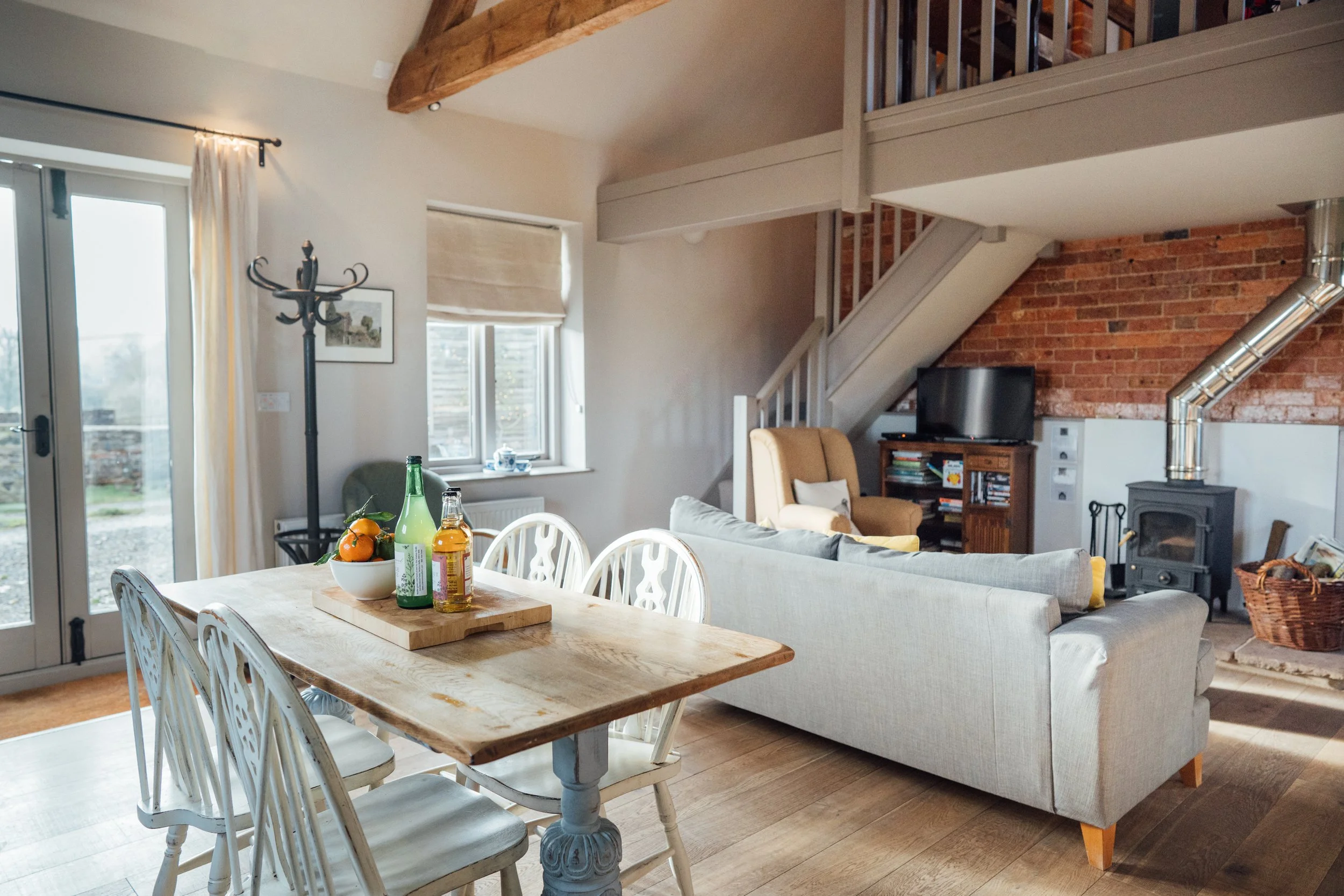 Living room with wooden dining table, chairs, light-colored sofa, brick wall, television, wood stove, and staircase.