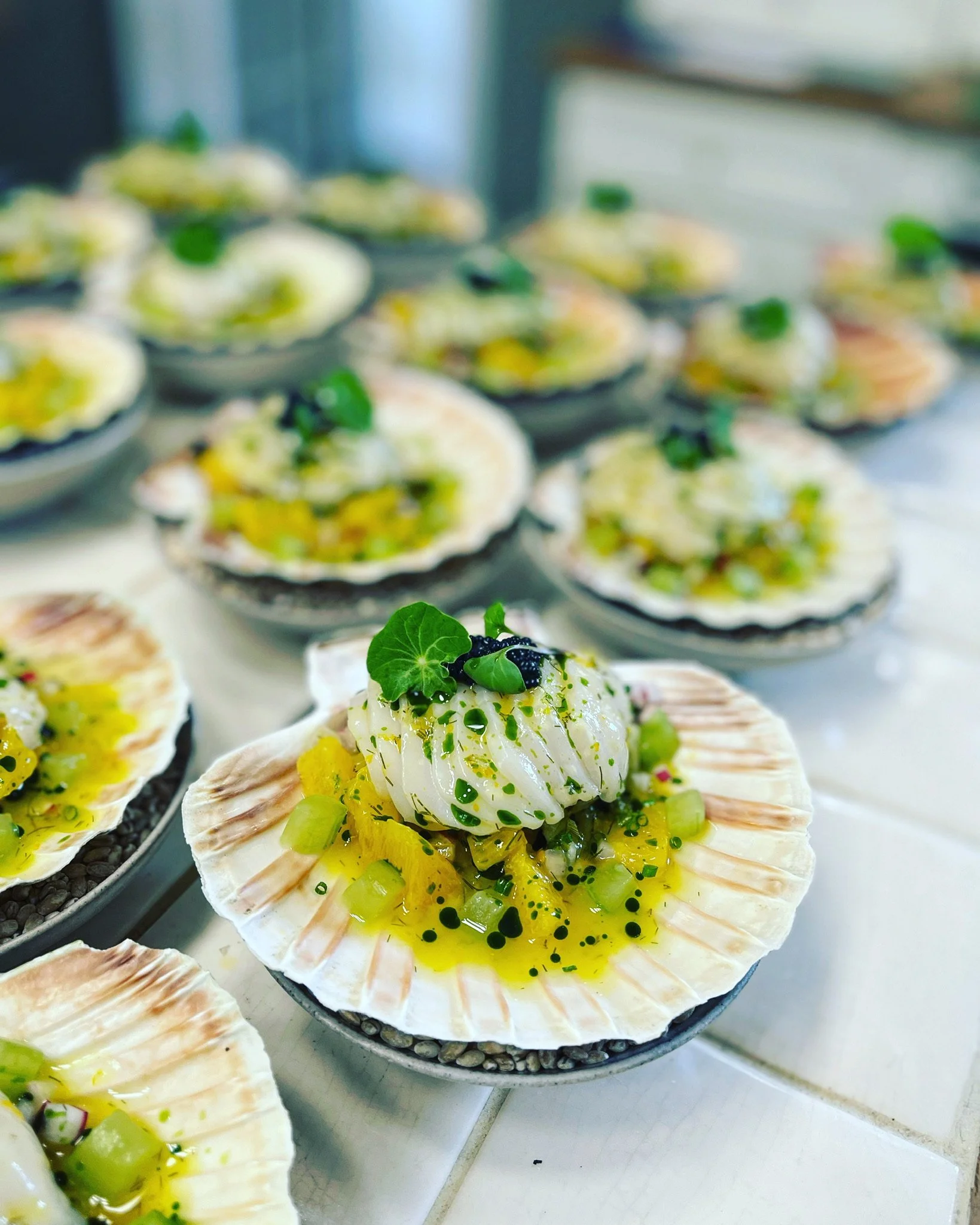 A plated scallop on a shell with green garnish and lemon slices, surrounded by similar dishes in the background.