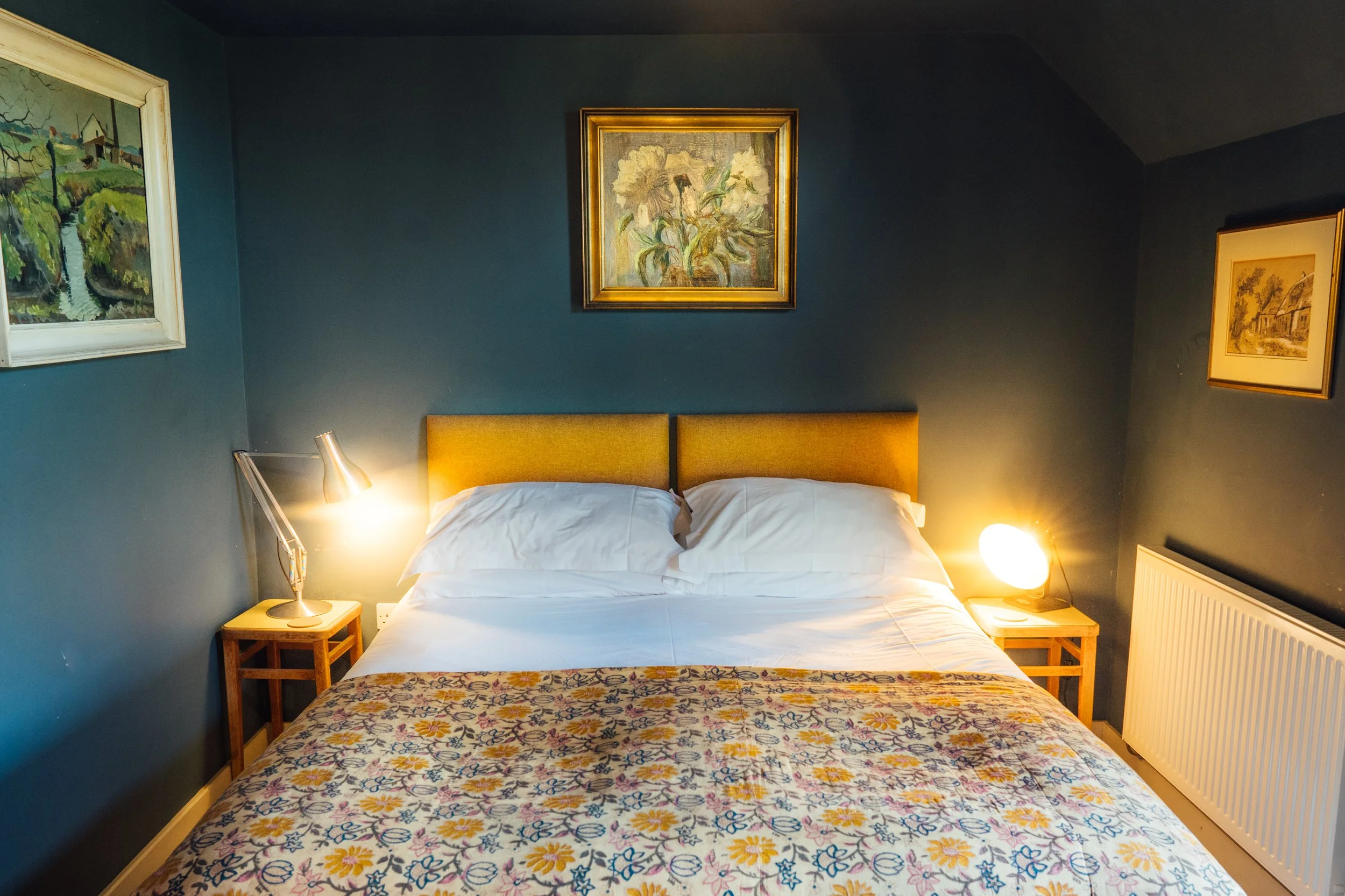A neatly made bed with white sheets and pillows, a yellow headboard, and two side tables with lamps. The room has a blue wall with three framed paintings above the bed and on the sides.