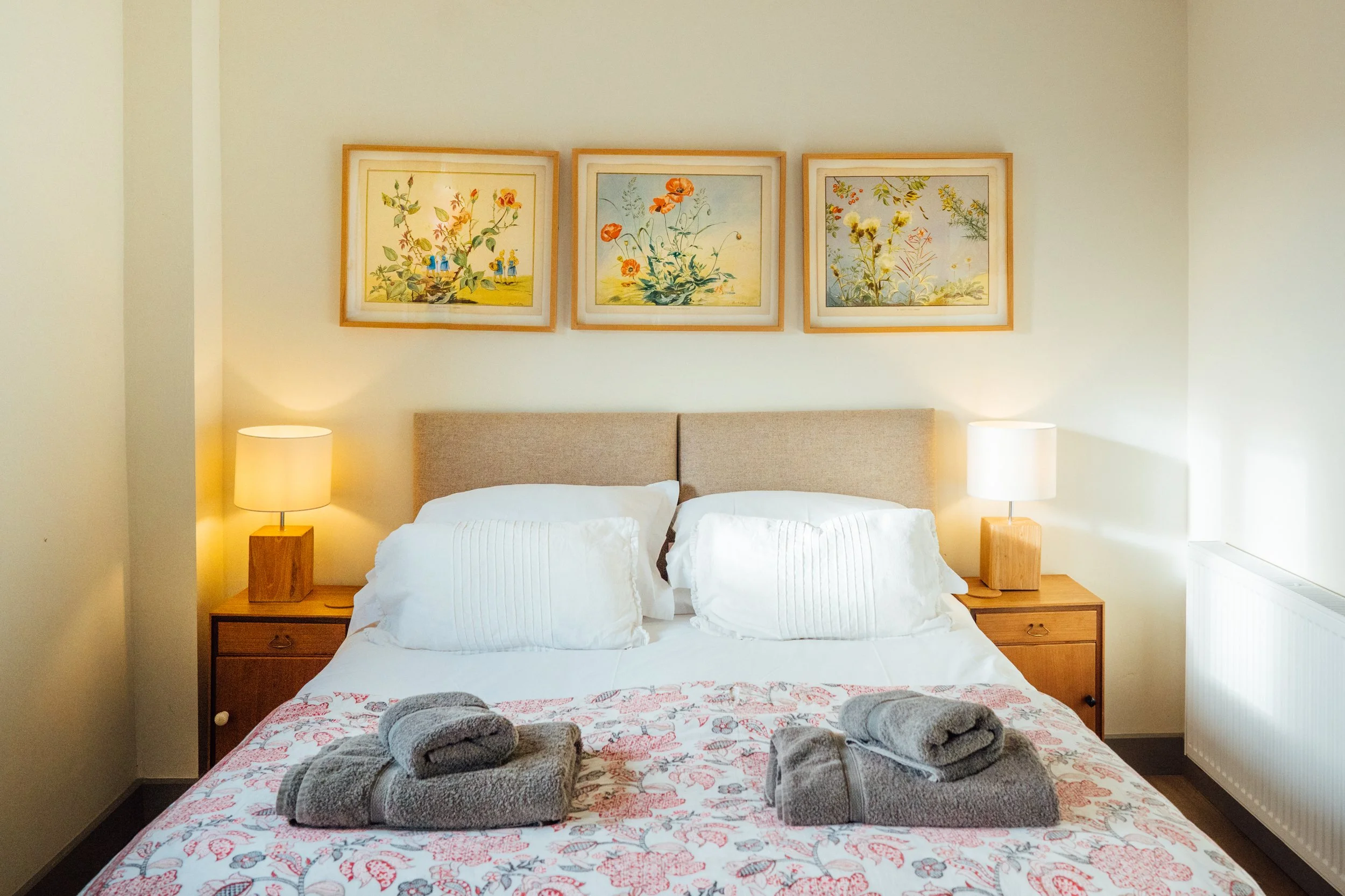 A neatly made bed with white pillows and floral bedspread, two matching gray towels on the bed, wooden nightstands with lamps, and framed botanical prints above the headboard.
