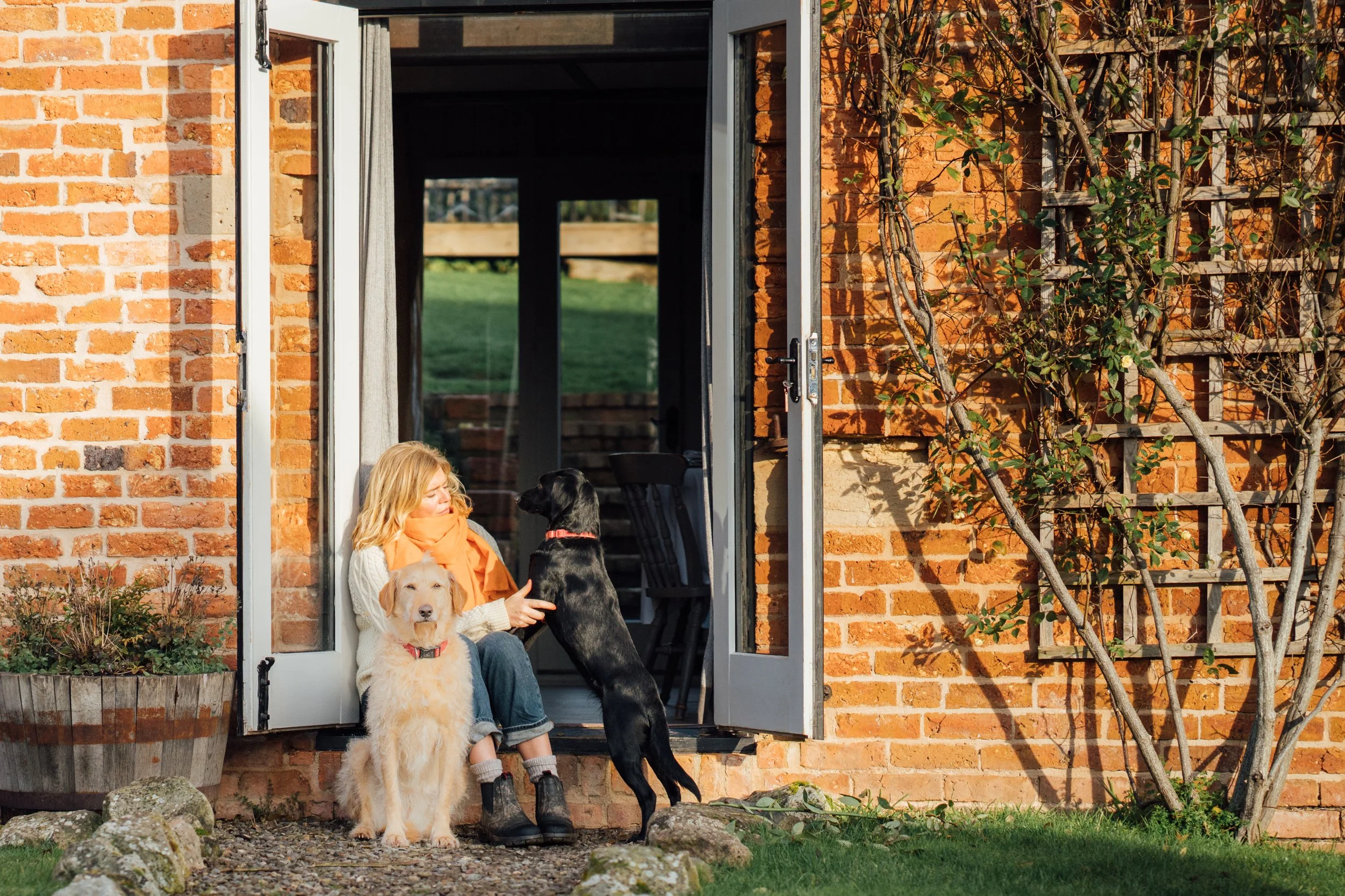 A woman with blonde hair sitting at her doorway with two dogs, one black and one yellow, during the daytime. The woman is reaching out to the black dog who is standing on its hind legs. The yellow dog is sitting close to her, facing forward. The hous