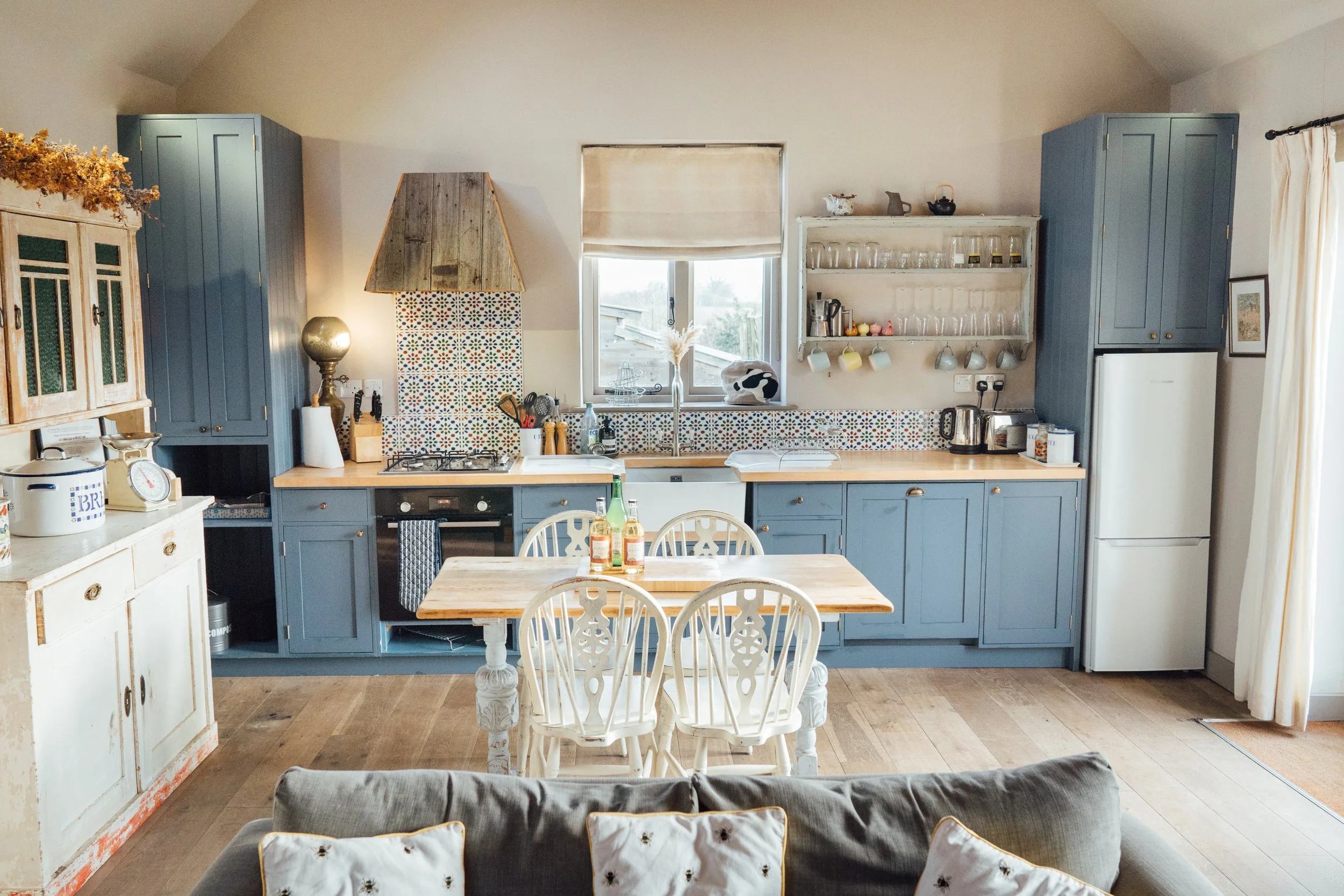 A cozy kitchen with blue cabinetry, a wooden dining table, white chairs, and a mix of vintage and modern decor in a bright, airy space.