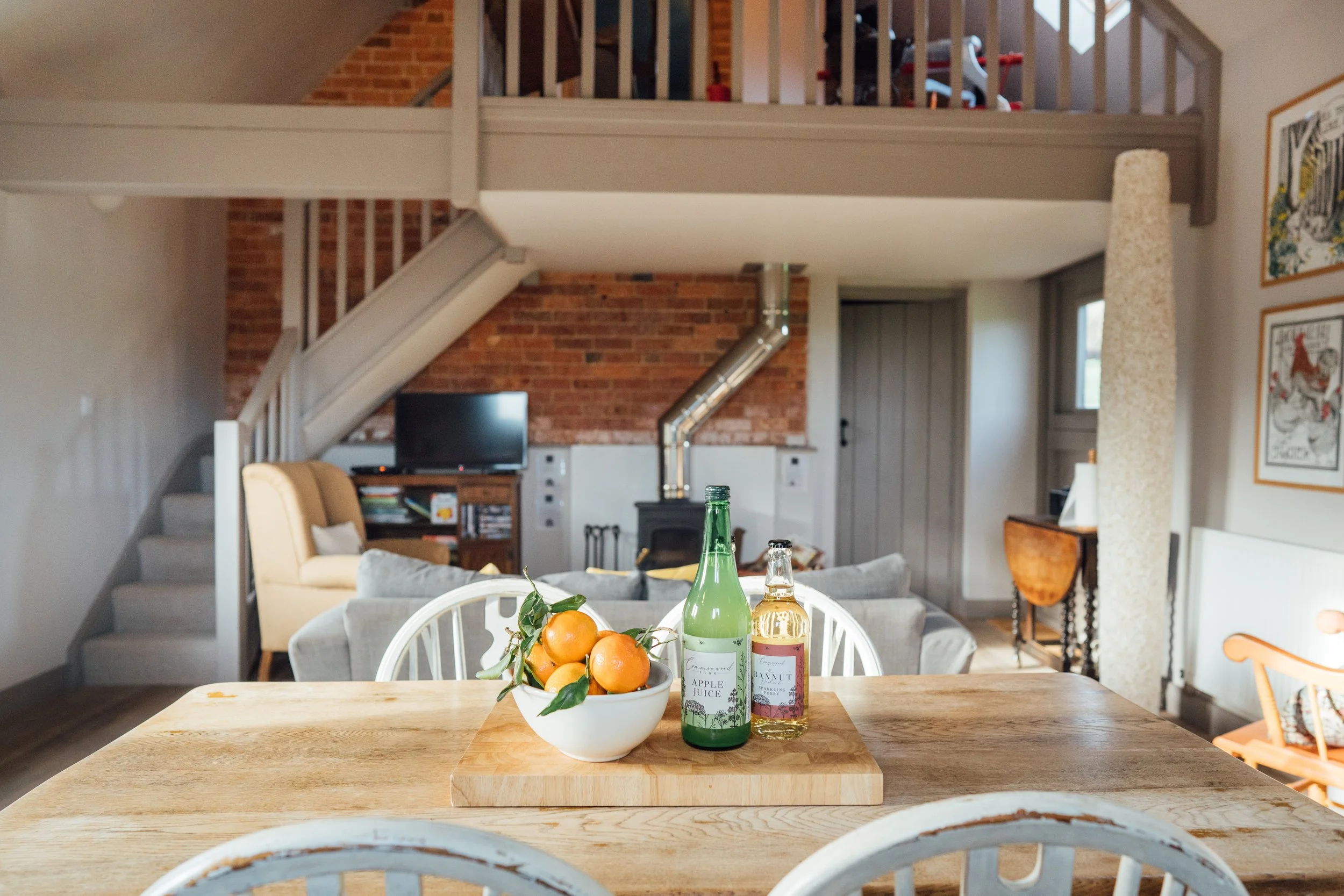 Kitchen table with a bowl of oranges and bottles of apple juice and nut liqueur in the foreground. Living room with a beige armchair, gray sofa, television, and fireplace in the background, with a loft area above.